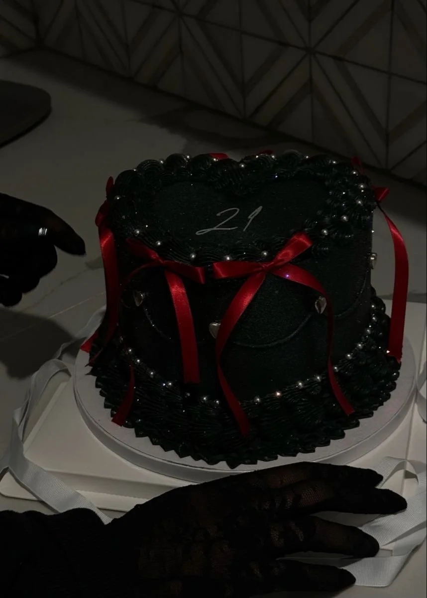 A black birthday cake with decorative piping, silver pearls, and red ribbons, featuring the number 21 on top, placed on a white cake board. Two hands in black gloves are near the cake.