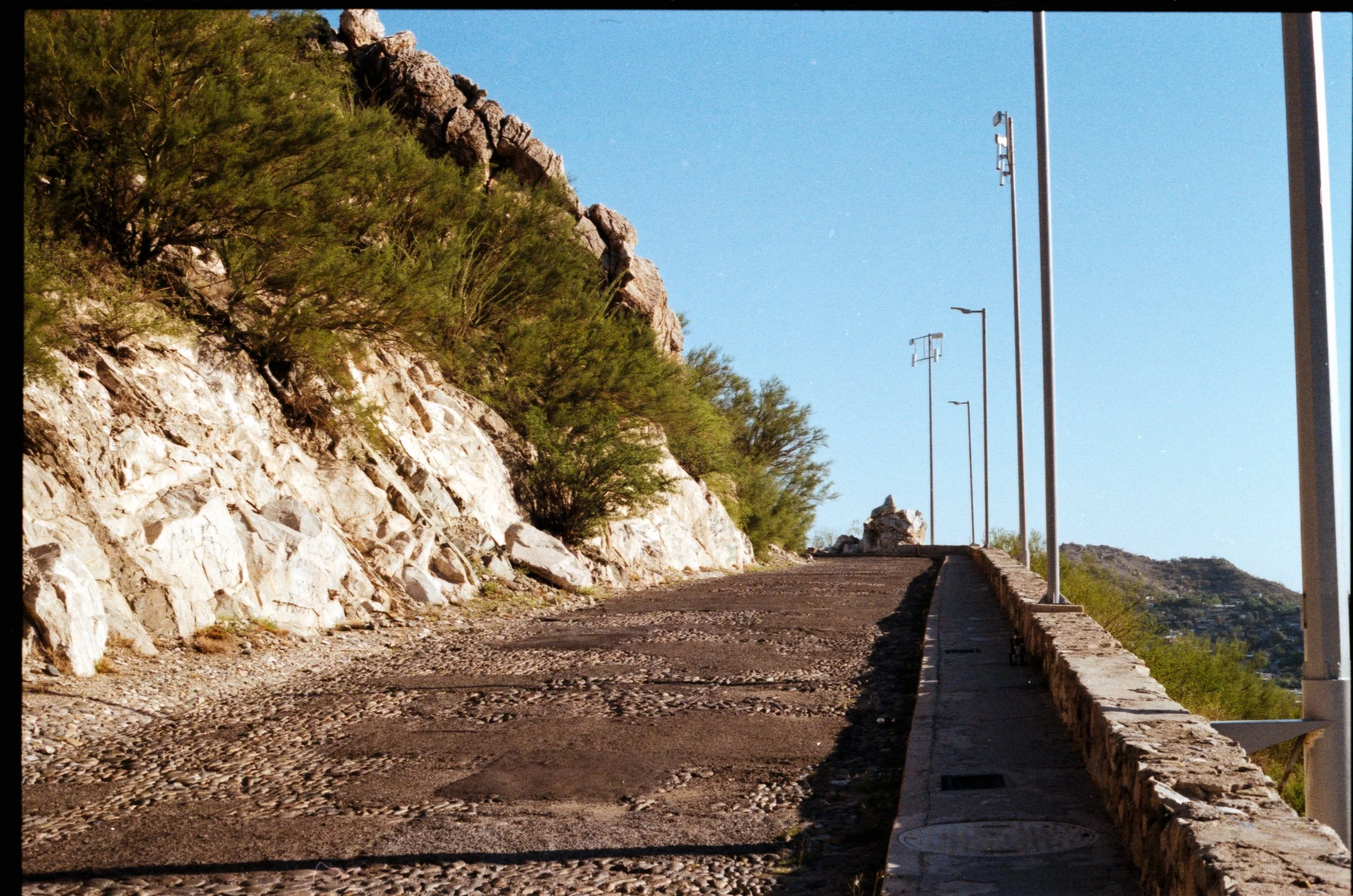 Camino ascendente junto a colina rocosa y vegetación, con postes de luz y cableado, en un día despejado.