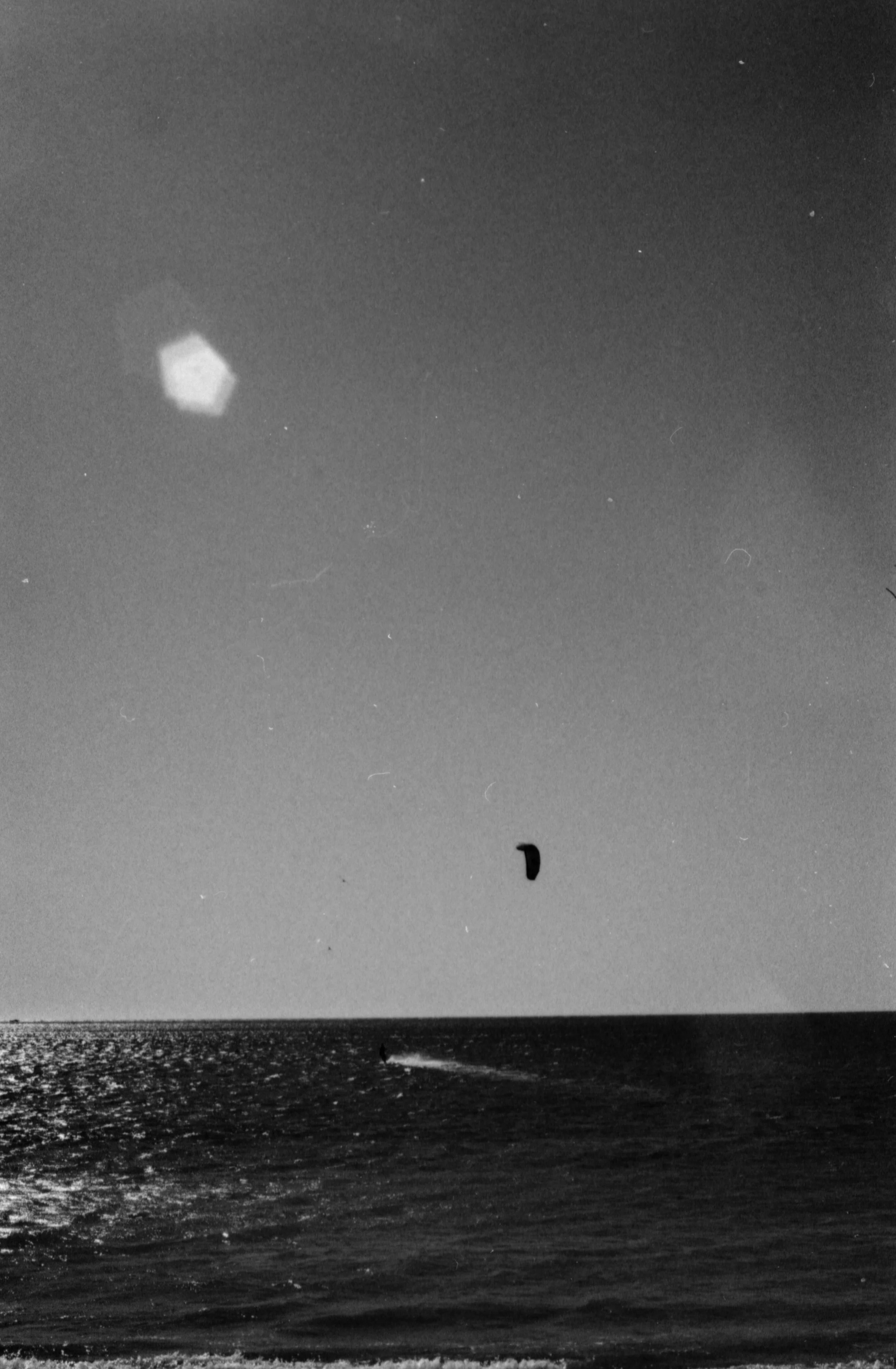 Vista en blanco y negro de un mar con un parapente en el cielo y la luna visible en la noche.