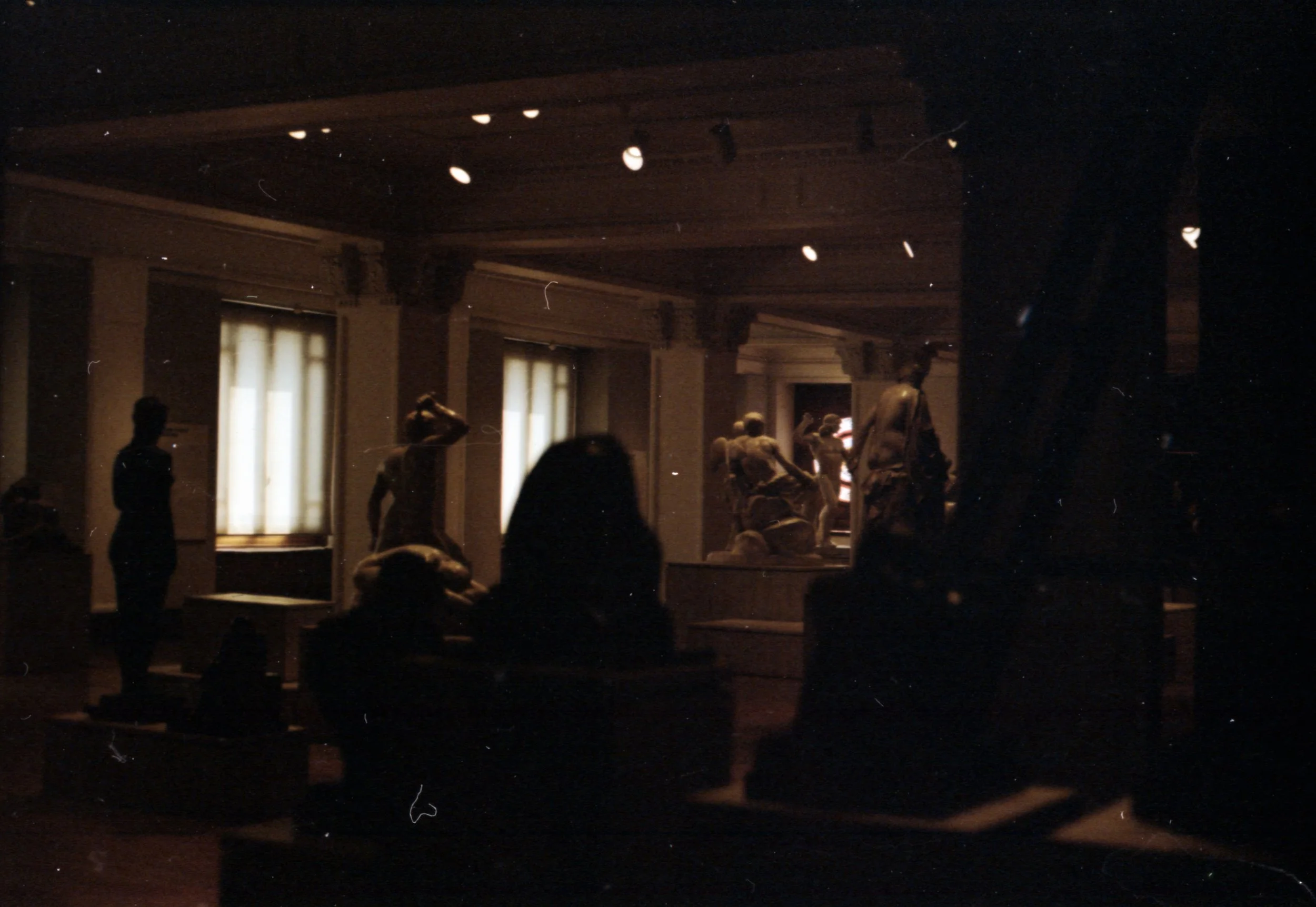 Esculturas en una exhibición en un espacio con poca iluminación y cortinas blancas en las ventanas.