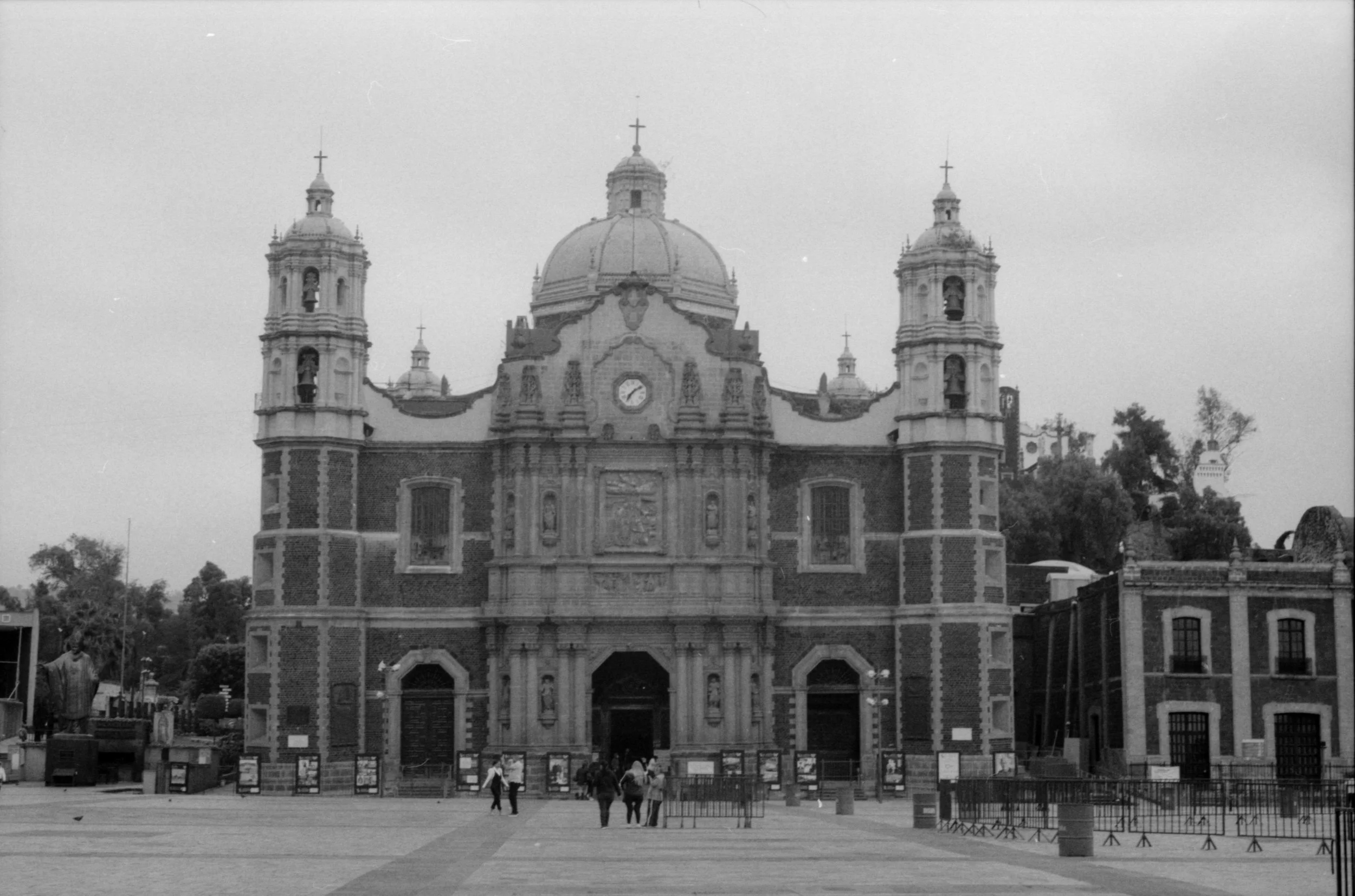 Una iglesia con arquitectura barroca, con doble torre y una cúpula central en un área abierta con varias personas caminando y árboles al fondo, en una ciudad de México.