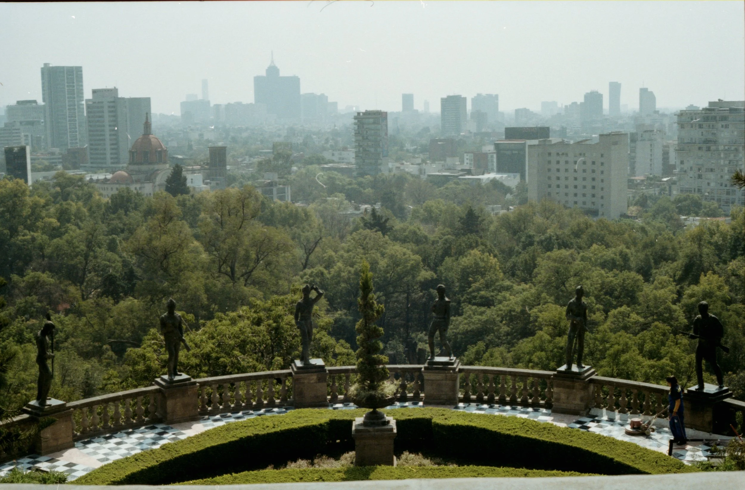 Vista panorámica de la ciudad desde un jardín con estatuas de personas en la barandilla y árboles verdes, en un día ligeramente nublado.