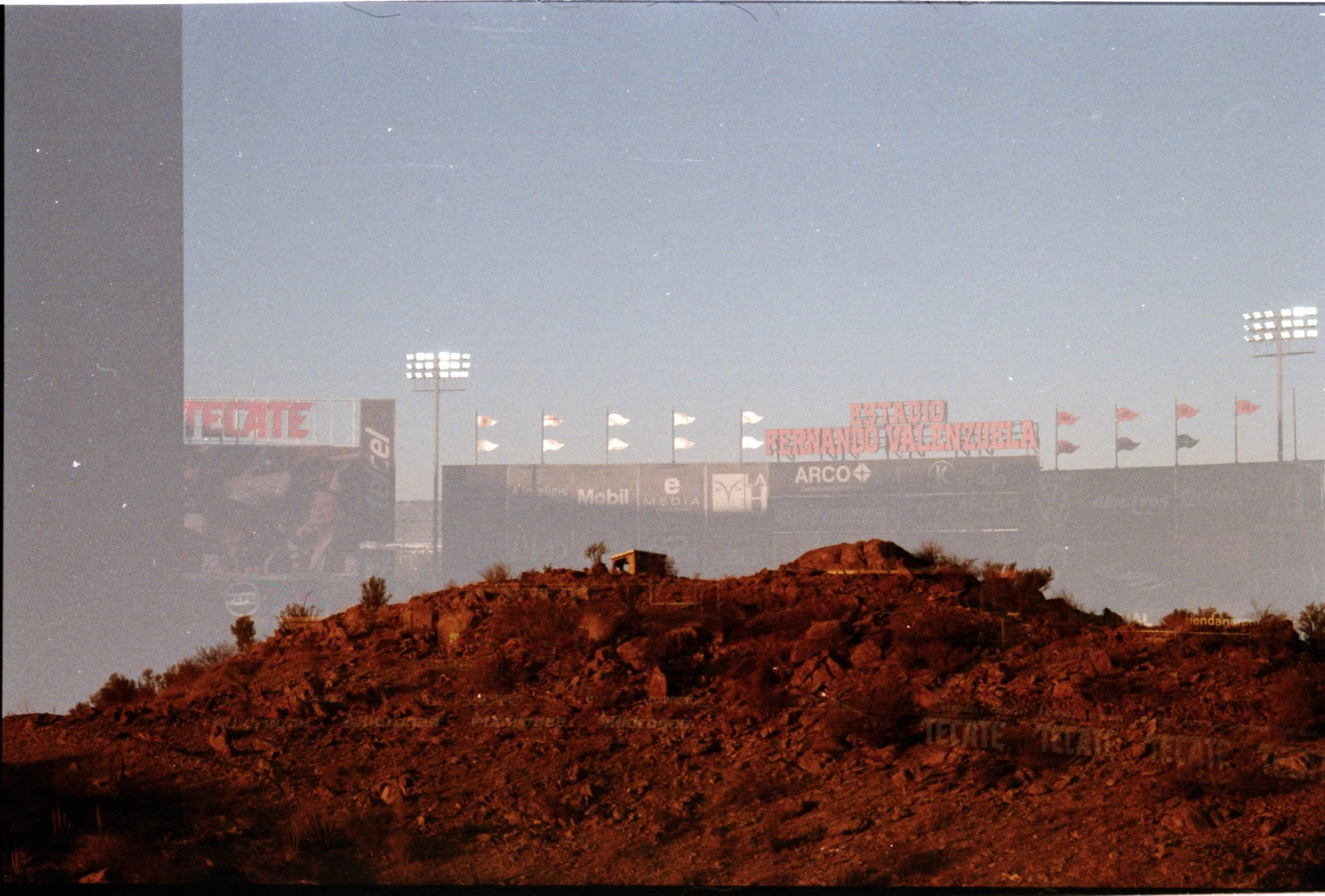 Imagen con doble exposición, mostrando un cerro con vegetación y un campo de fútbol en la cima, con cartel que dice 'Estadio Fernando Valenzuela' y cartelera con anuncios publicitarios.
