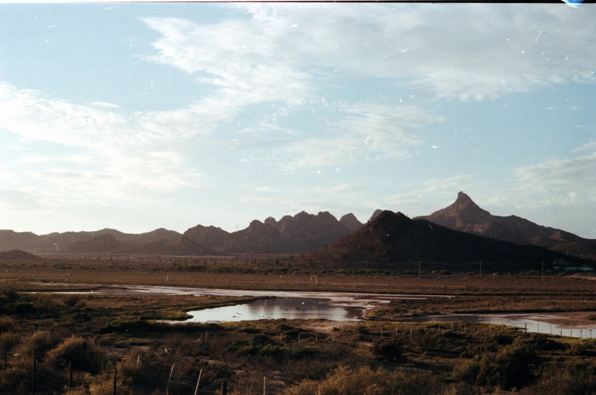 Paisaje árido con montañas rocosas y un río de agua tranquila en primer plano, cielo parcialmente cubierto de nubes.