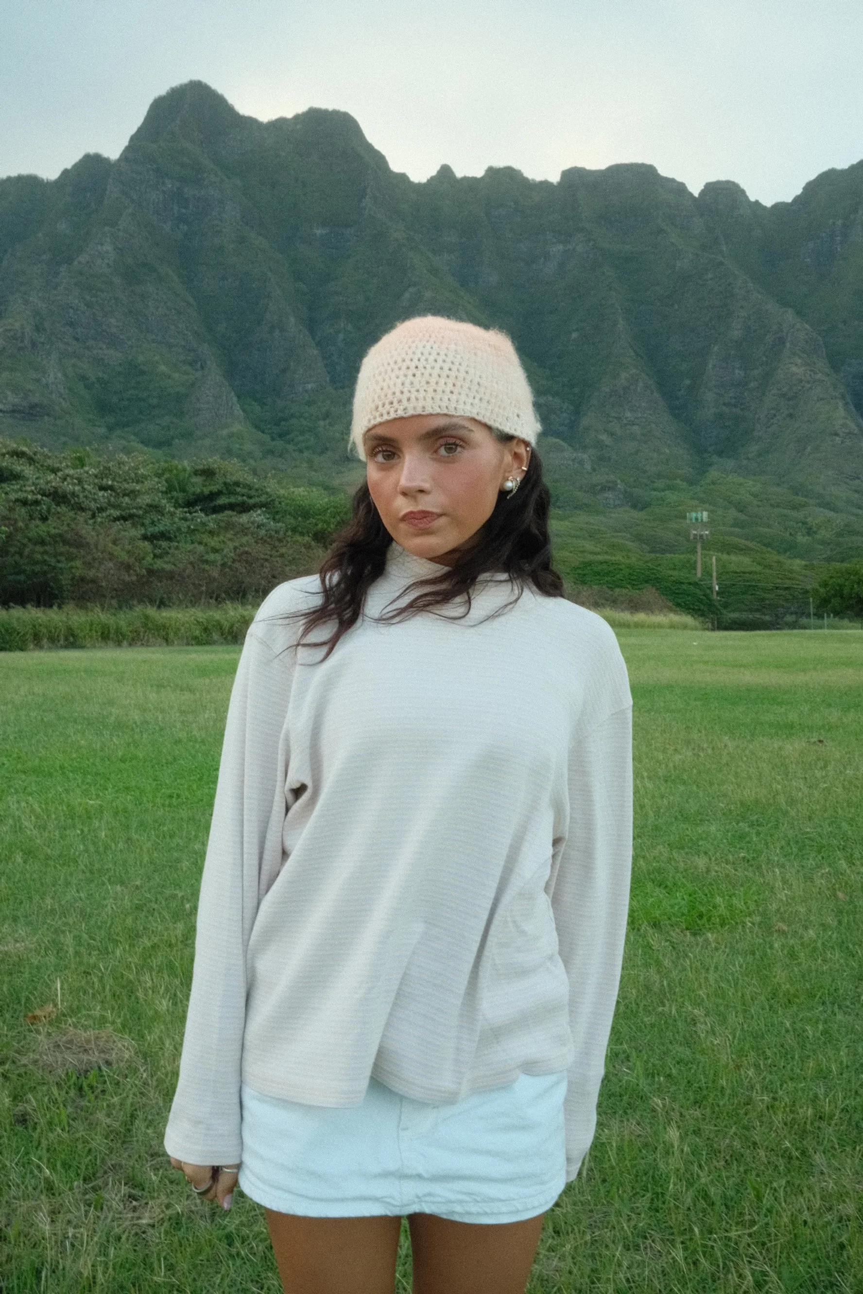 Young woman standing on a grassy field with green mountains in the background, wearing a beige knit hat, a white long-sleeve shirt, and white shorts.