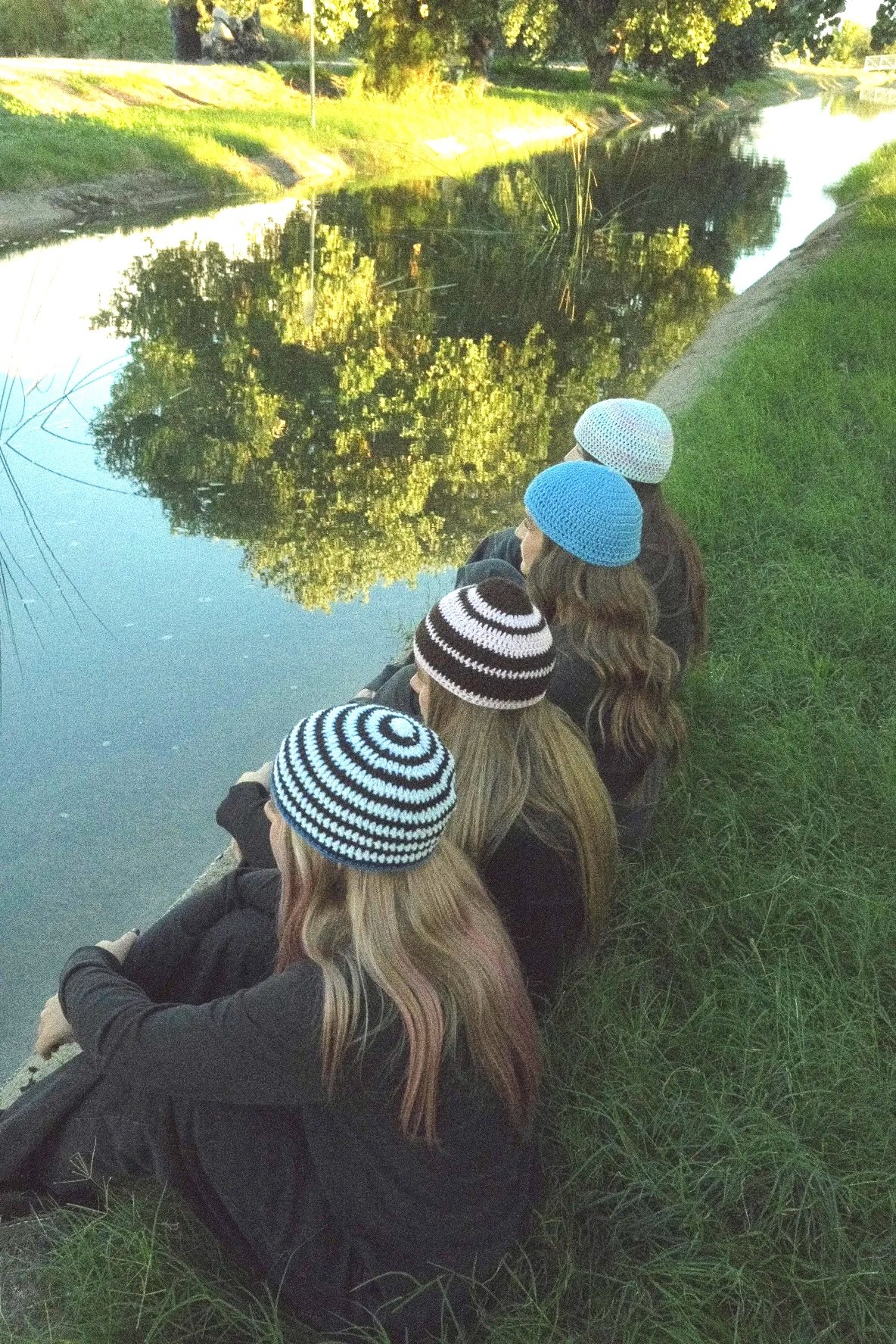 Five people sitting on the grass by a canal, wearing knit hats. The water is calm with reflections of trees, and the scene is illuminated by soft, natural evening light.