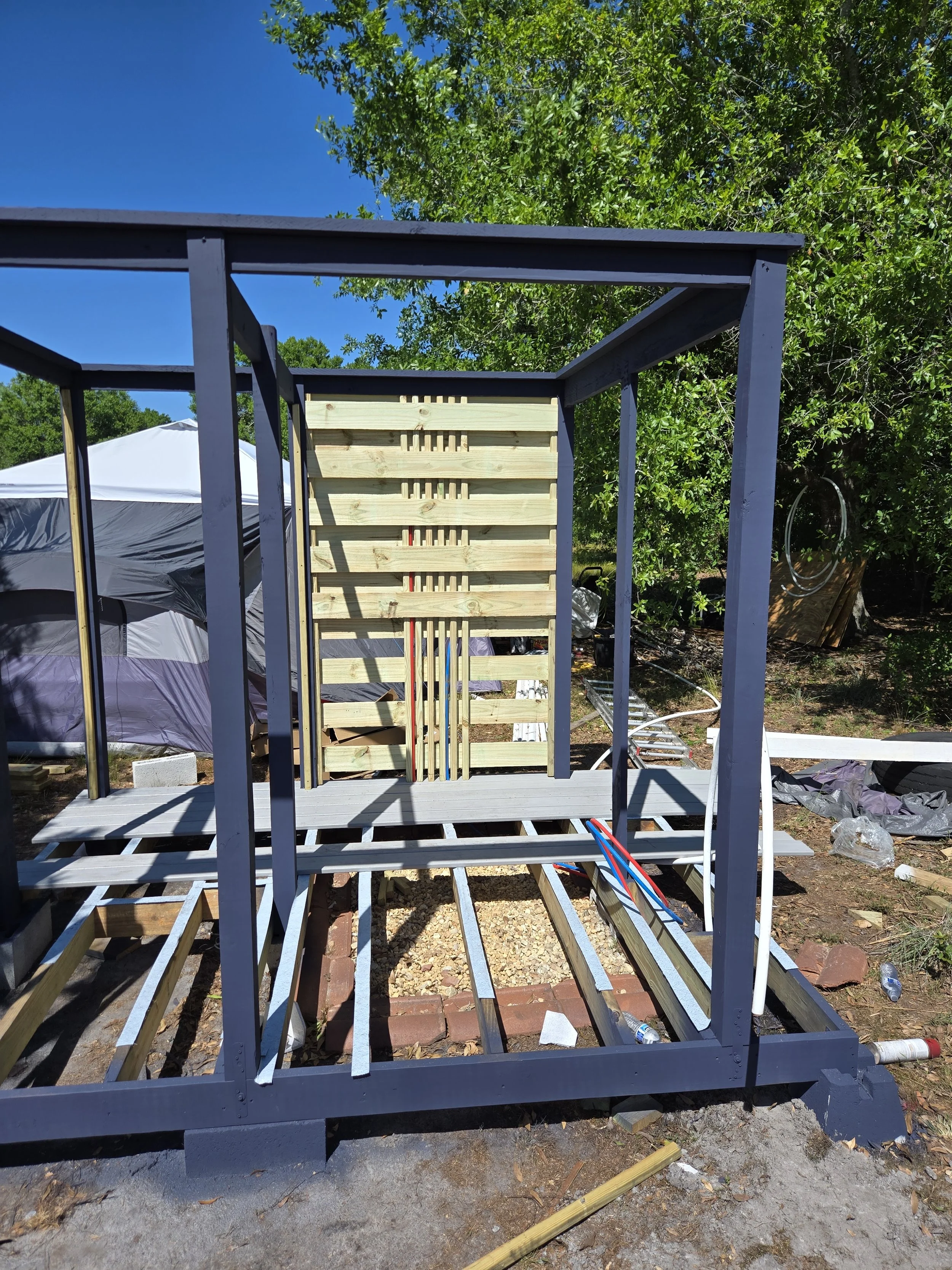 Construction of a wooden deck with a gray painted frame and a partially built wooden privacy wall, in an outdoor backyard with trees and a tent in the background.