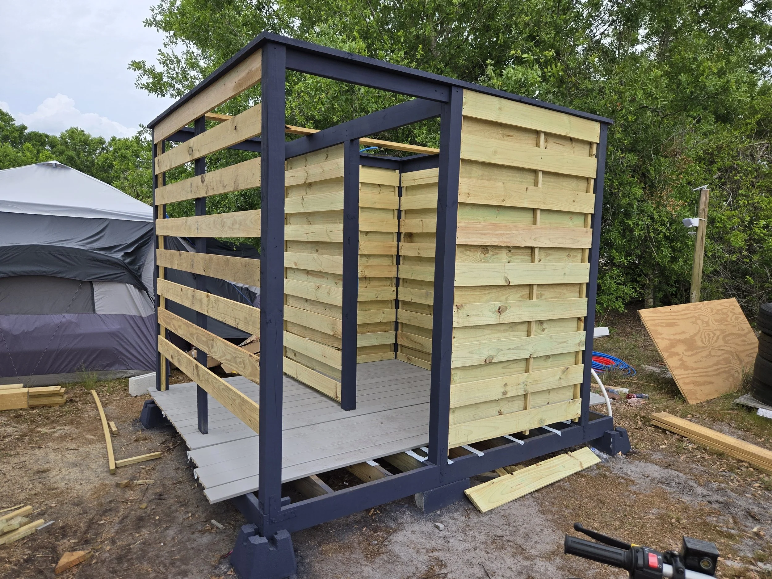 Wooden structure under construction on a deck, with some walls made of horizontal wooden planks and a partially built enclosure, outdoors with trees and construction materials around.