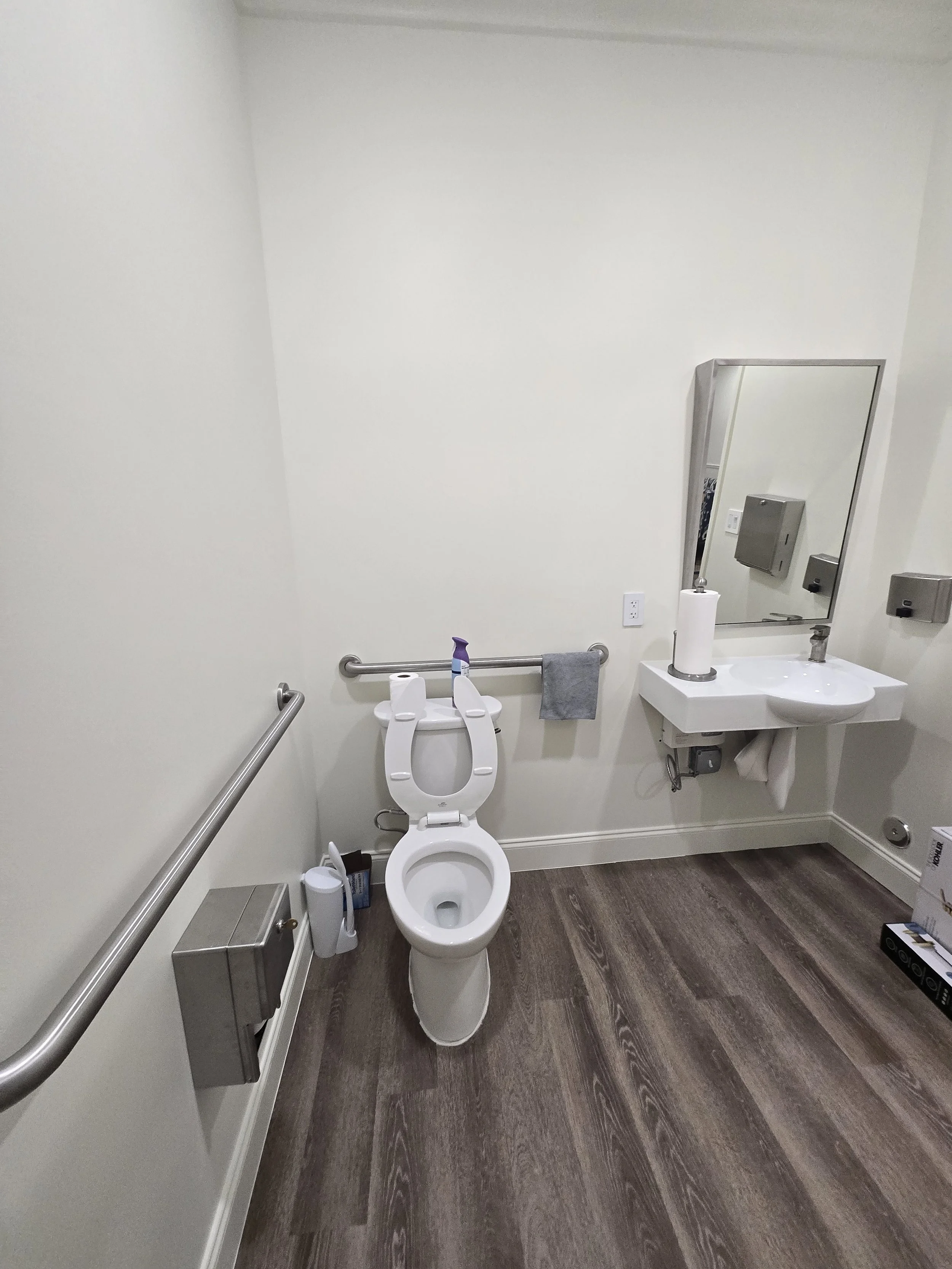 An accessible bathroom with a white toilet, a gray towel on a grab bar, a small white sink with a mirror, a paper towel roll, and various items on the floor and countertop.