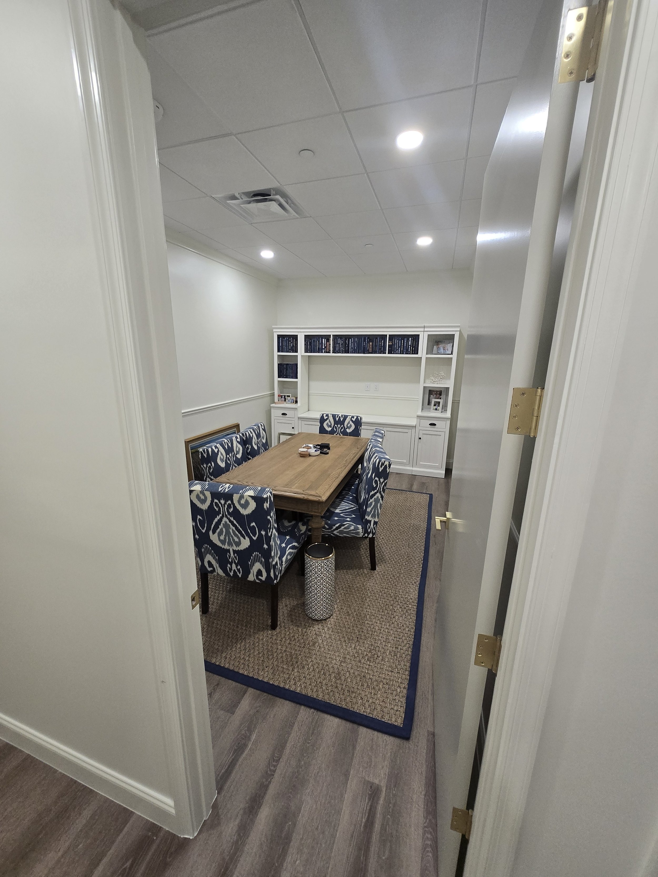 A small conference room with a wooden table surrounded by six blue and white patterned chairs, white built-in shelves at the back, ceiling lights, and dark wood flooring.