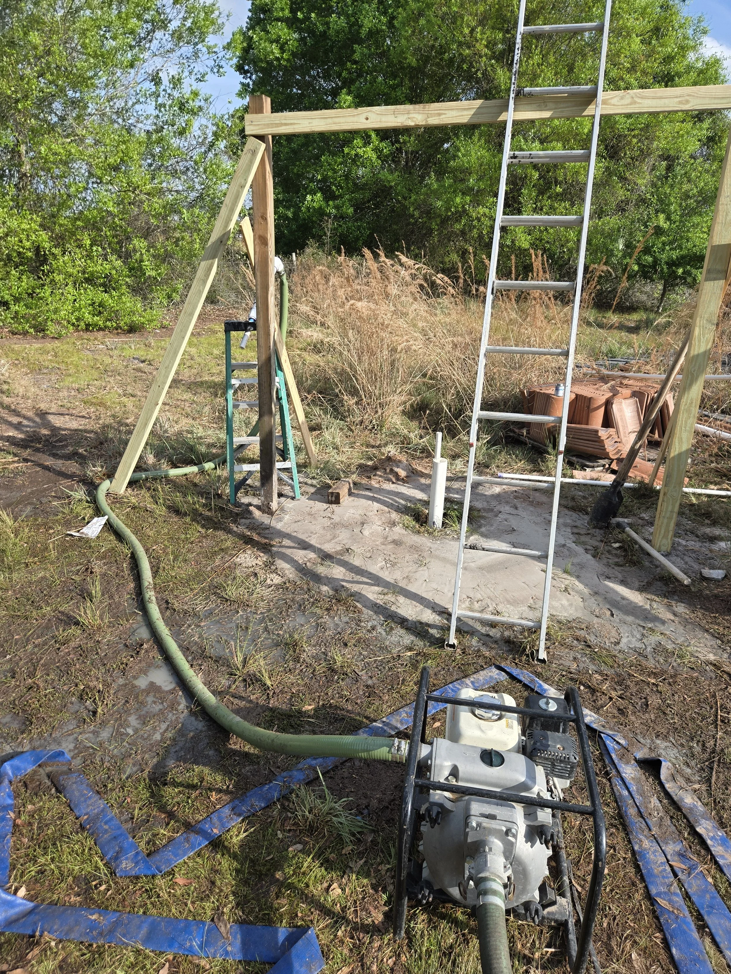 Well Construction site with wooden beams, ladders, and a portable pump on muddy ground surrounded by trees and construction materials.