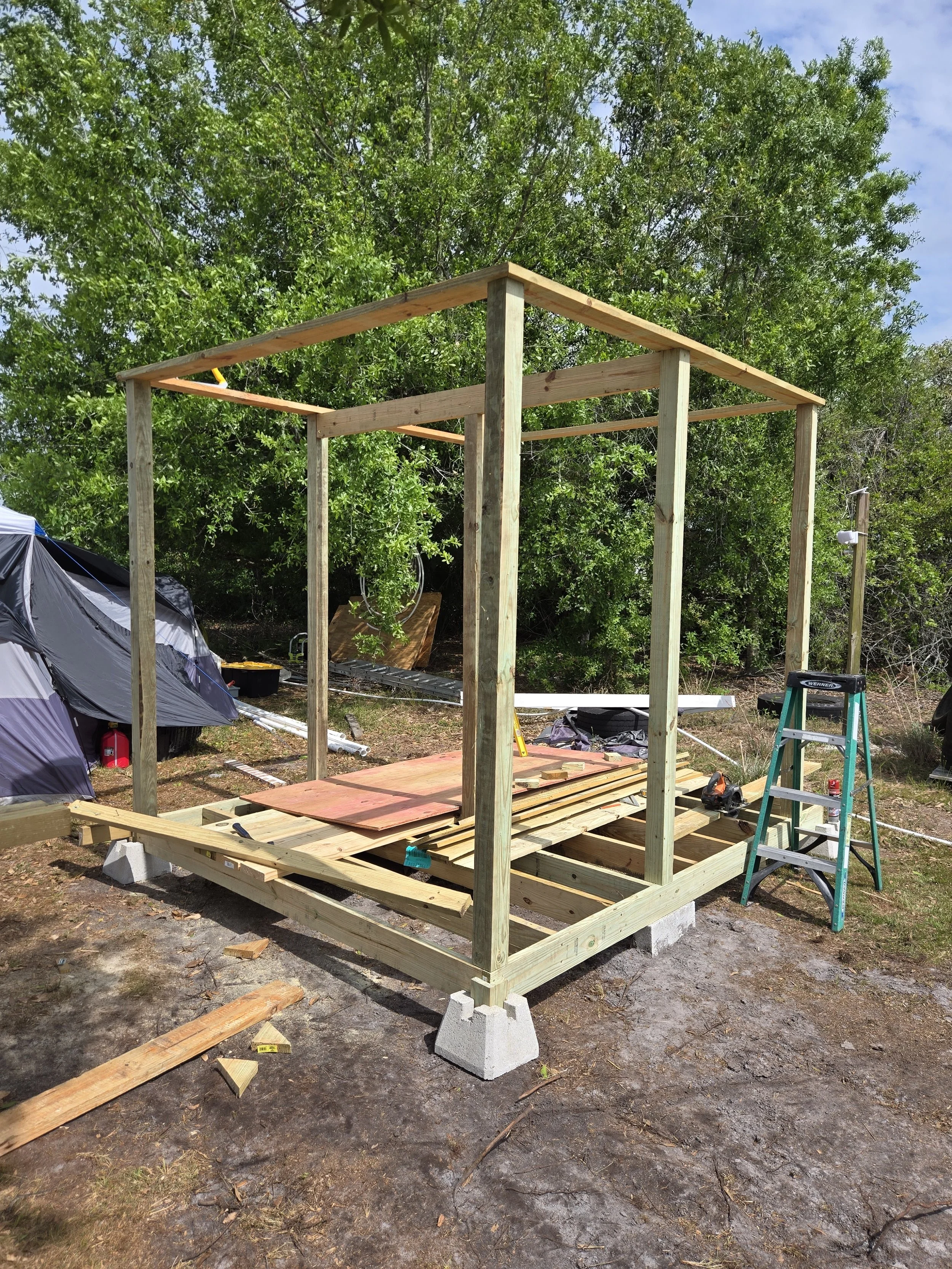 Wooden outdoor shower under construction with four vertical posts on concrete blocks, working tools, and building materials on a partially built wooden foundation.