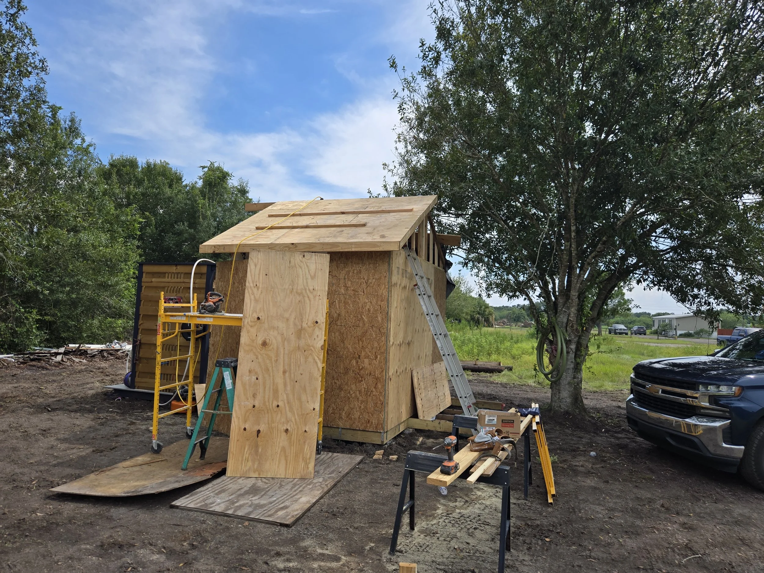 A small wooden shed under construction outdoors, with plywood and framing work in progress, surrounded by trees and parked vehicles.