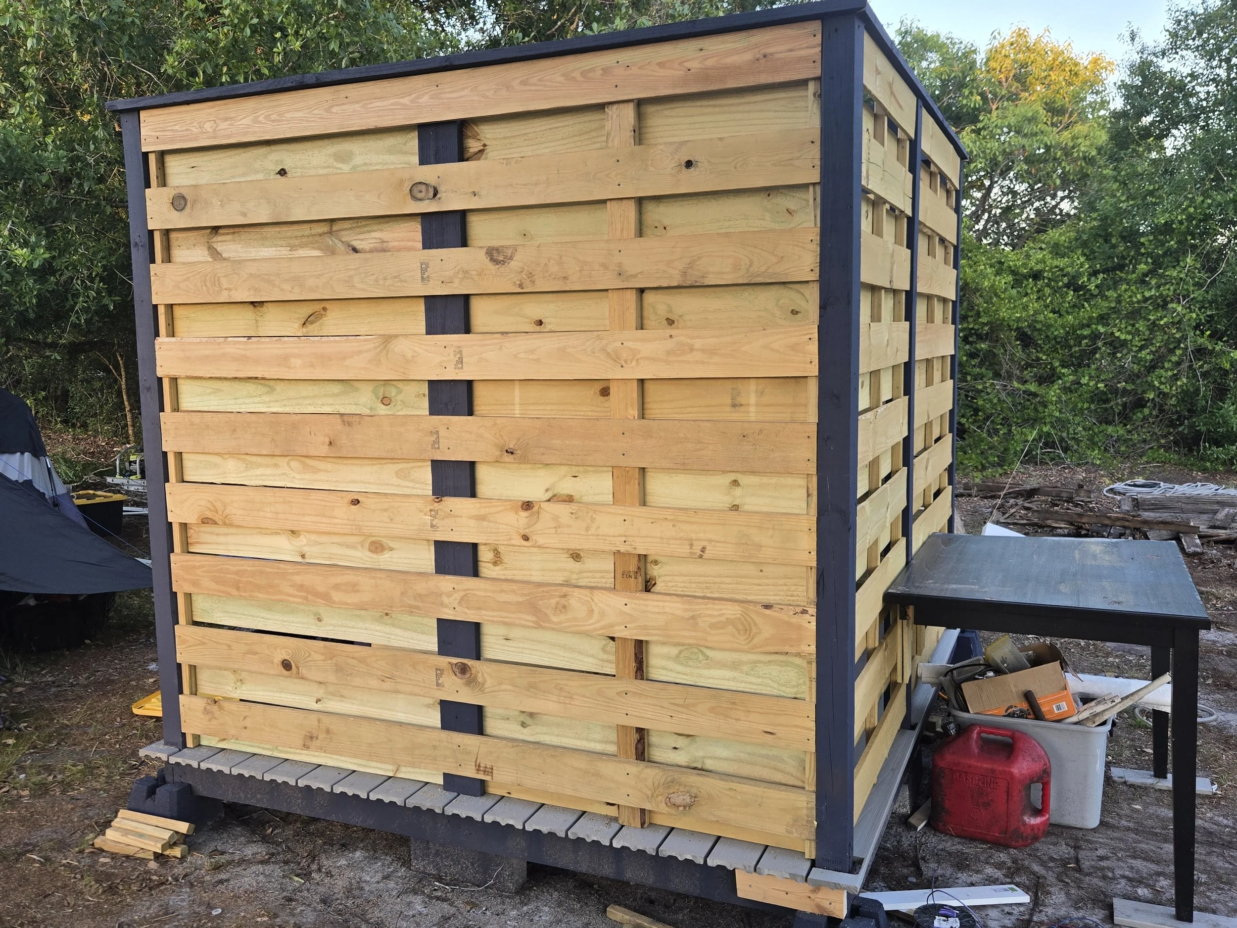A small wooden structure with horizontal slats in an outdoor setting surrounded by trees, with construction tools and supplies nearby.