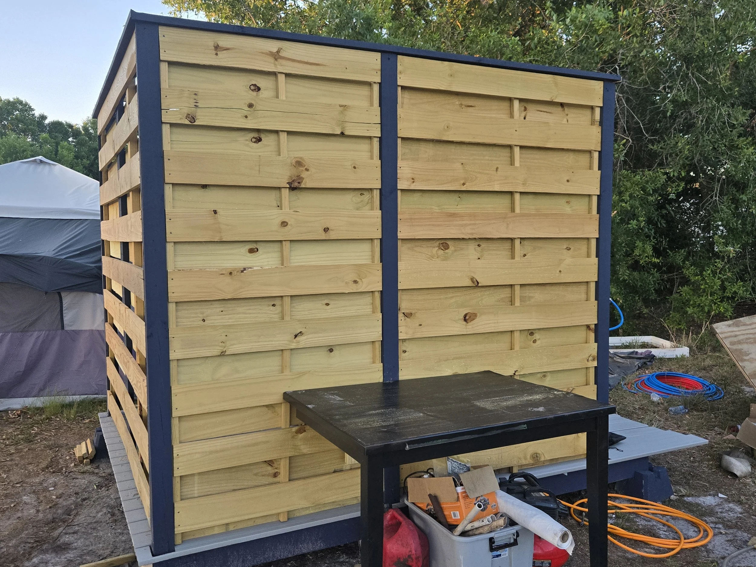 A small outdoor wooden shower with a blue frame, built with horizontal wooden planks, situated on a concrete base off grid.