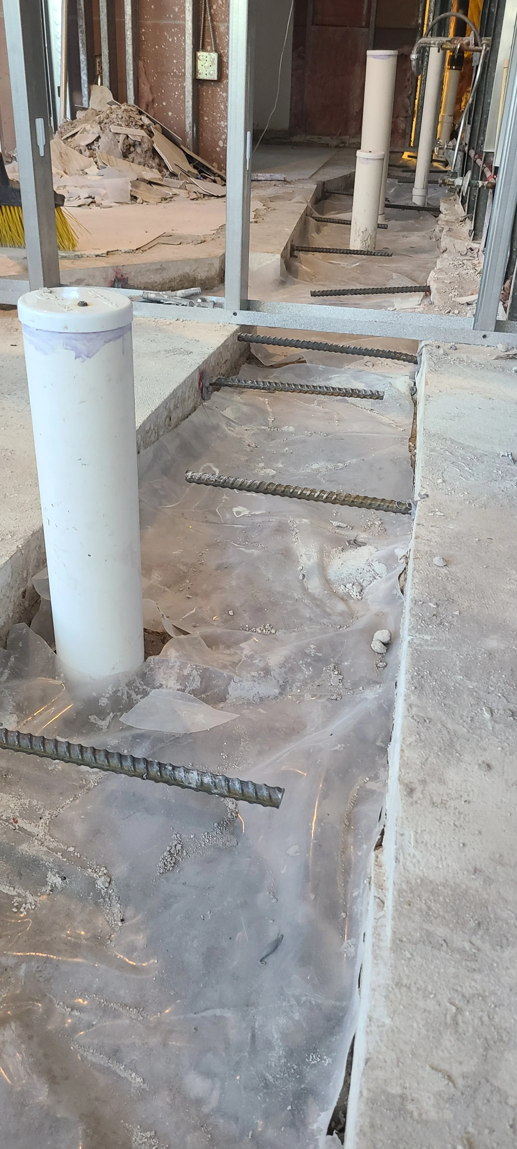 Construction site with partially built concrete or tile flooring, exposed rebar, and plumbing pipes. Piles of debris and construction materials are visible in the background.