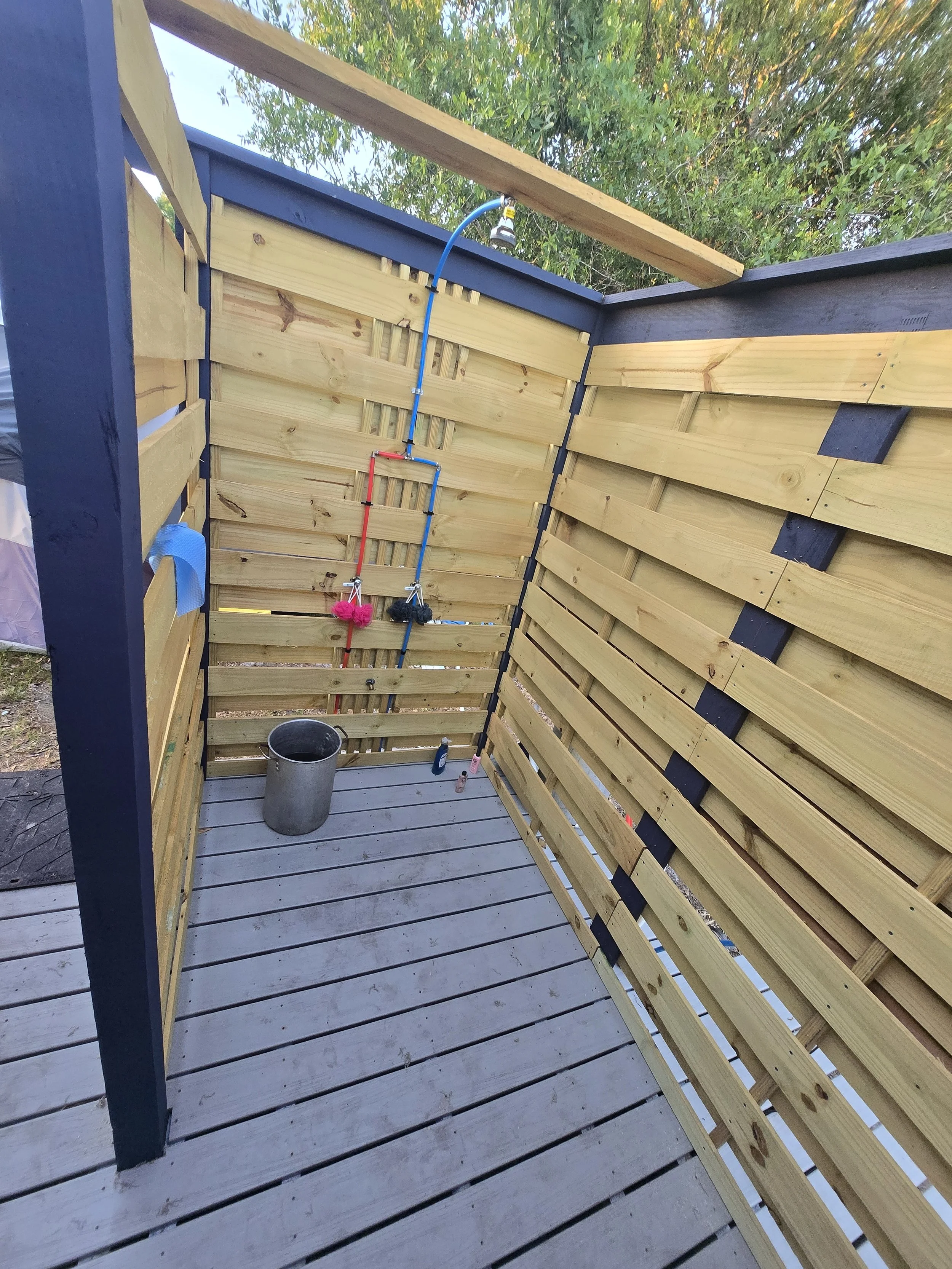 Outdoor shower station with wooden walls, a outside shower head connected by blue and red inlet pipes, fully usable water source to solar well.