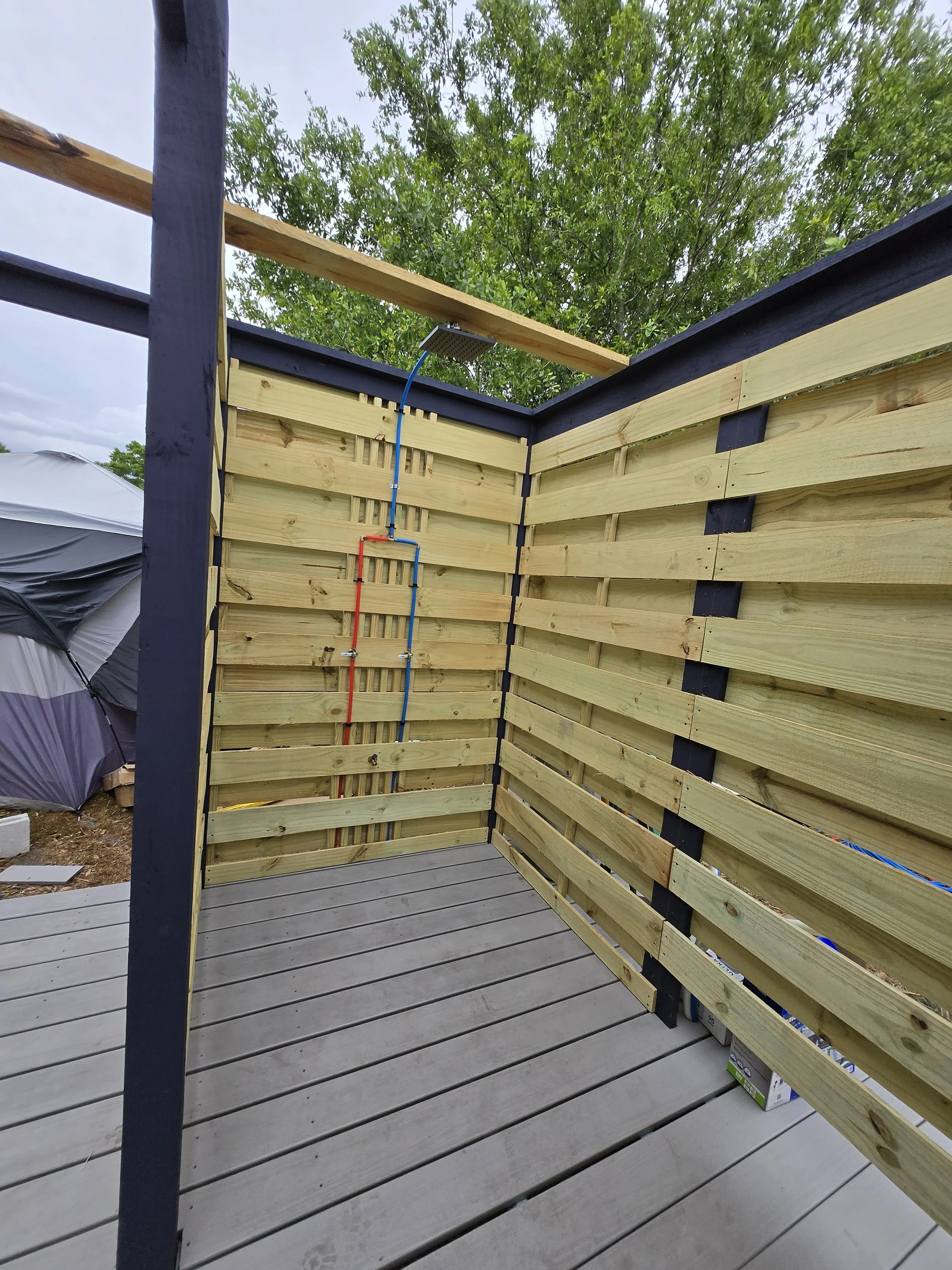 Construction of a shower on a wooden deck with unfinished wood walls, black trim, exposed plumbing, and an outdoor setting with a tree and tent in the background.