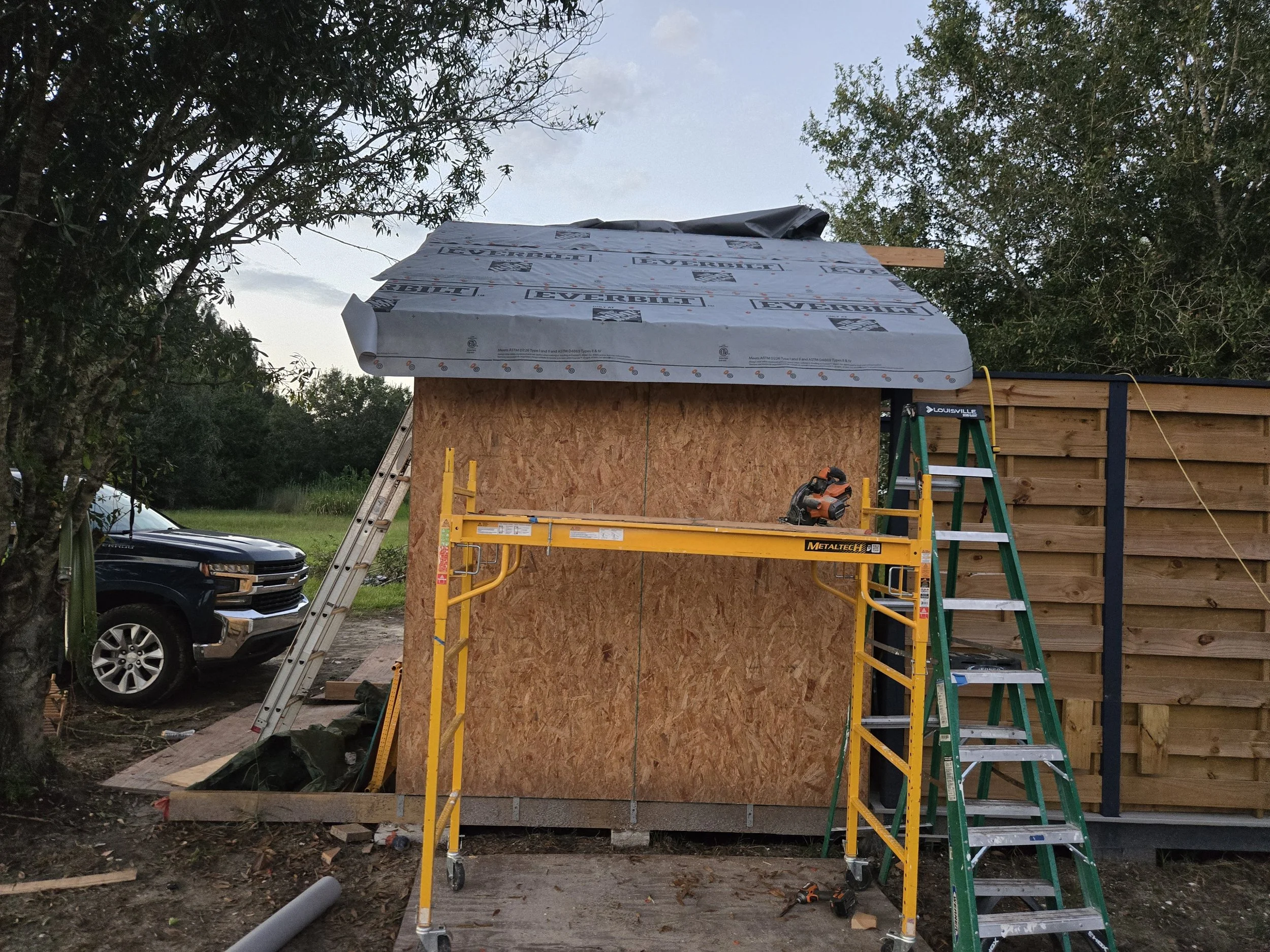 Construction site with plywood walls, a yellow scissor lift, and a green step ladder, with trees and a parked vehicle in the background.