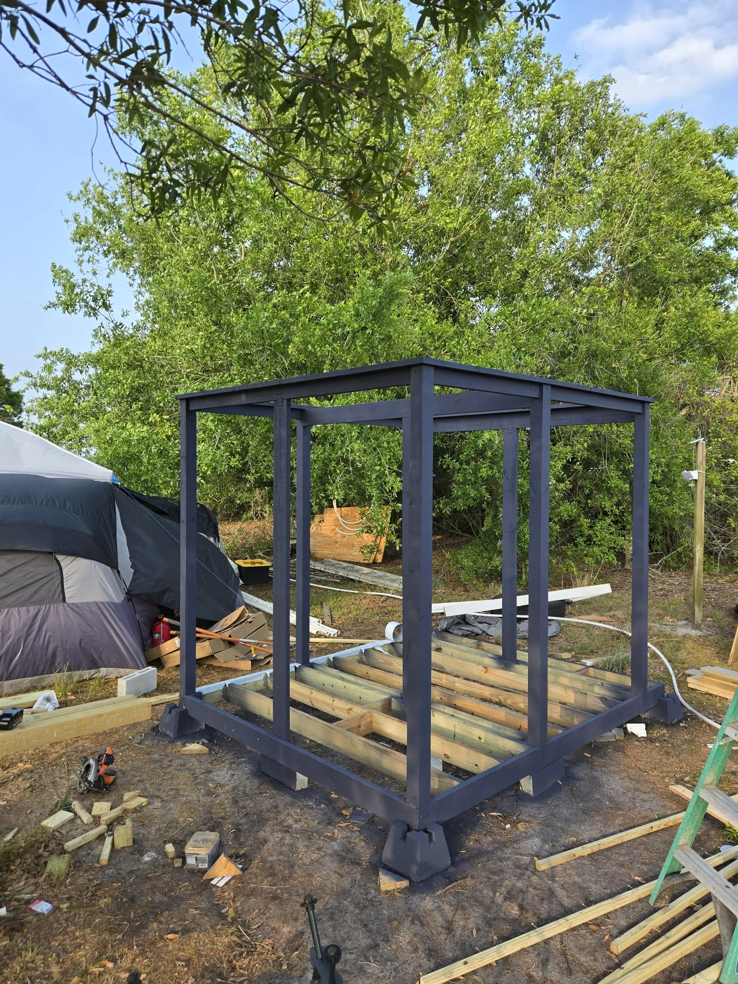 A metal frame structure under construction outdoors with lumber and construction tools scattered around, surrounded by trees and sky.