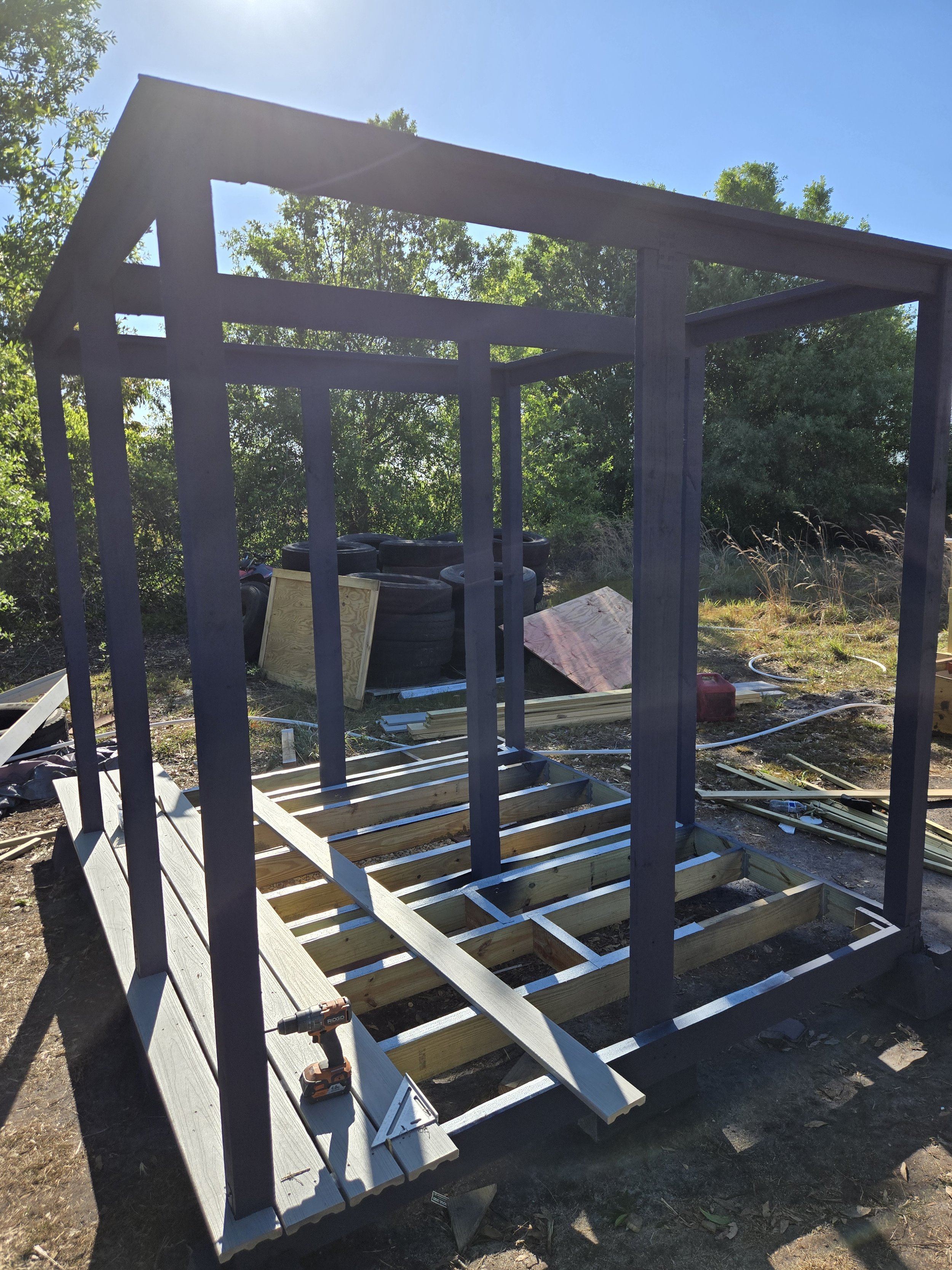 Construction of a small shed or playhouse with a wooden frame, outdoors in a backyard, with building materials and tires in the background.