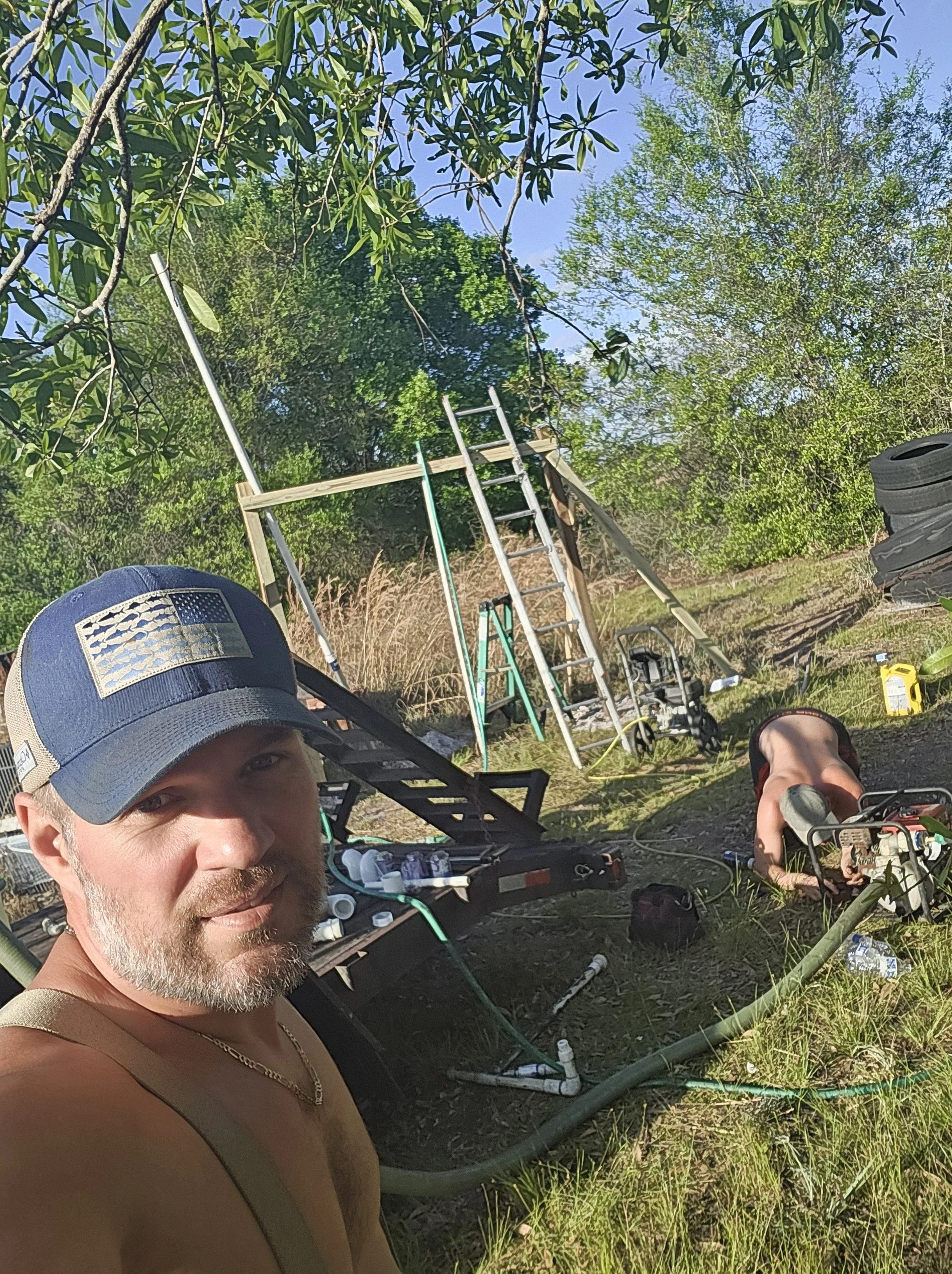 Owner selfie outdoors with a ladder and man working on well pump equipment in the background, prepping for water bore.