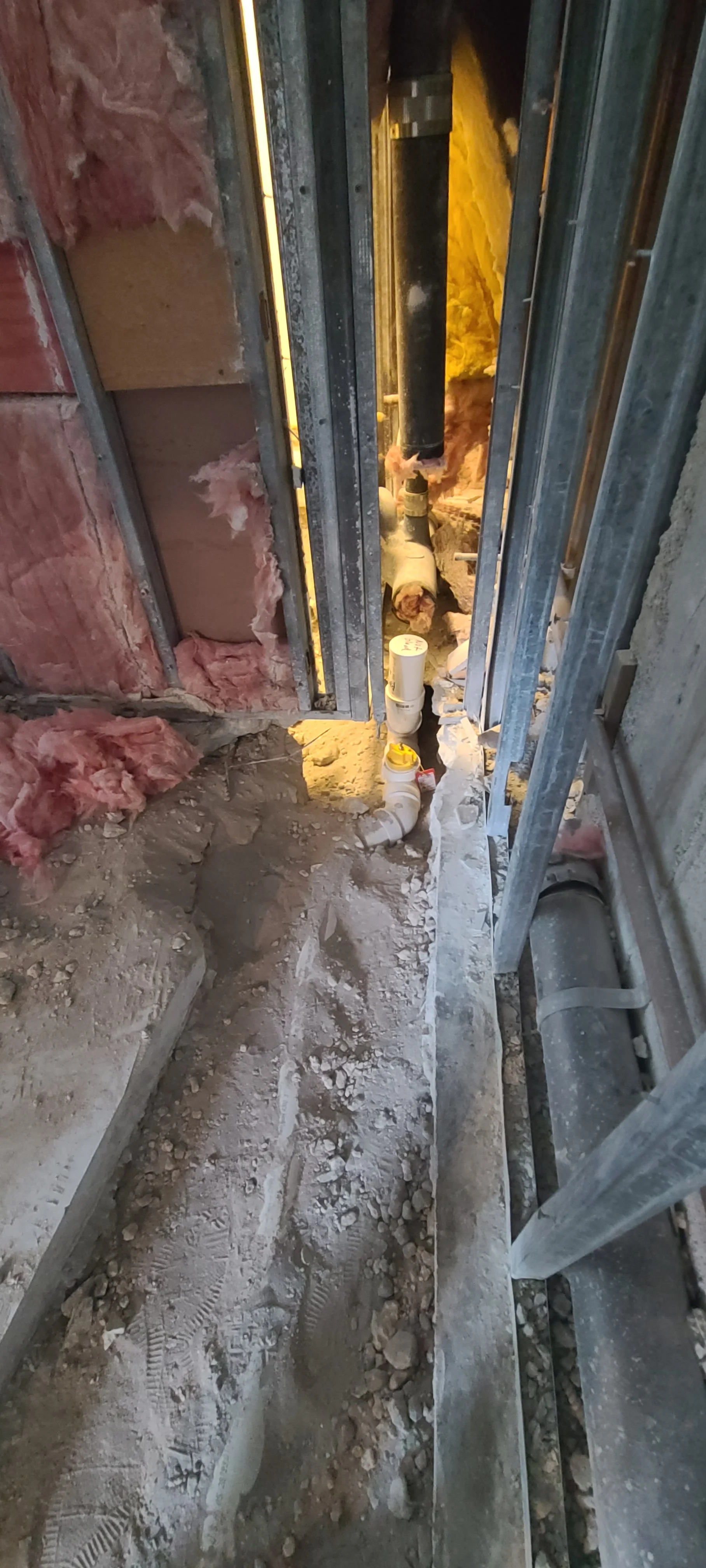 Under construction site showing exposed pipes, insulation, metal framing, and a dirt floor.