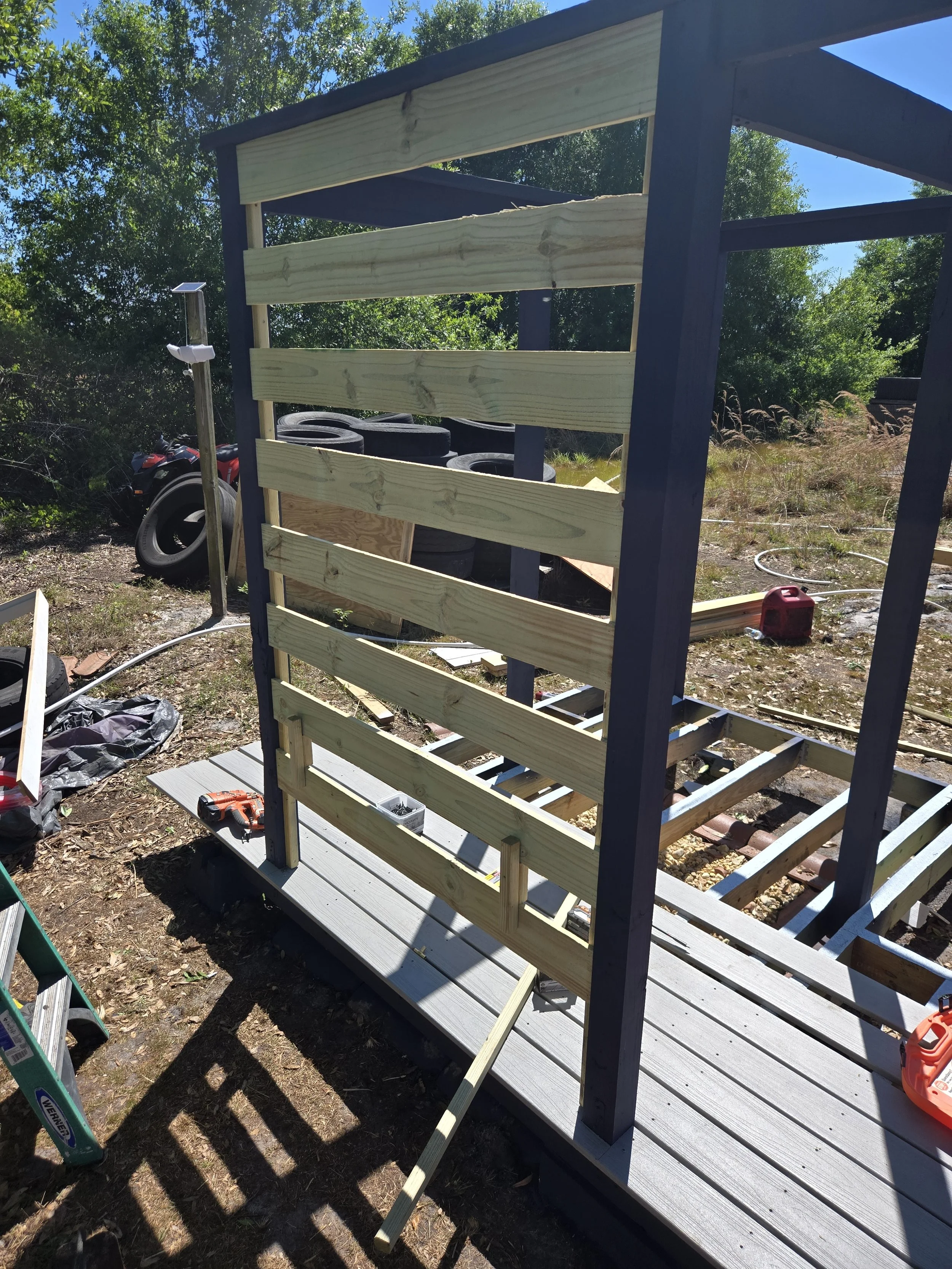 Construction of a wooden deck or porch with a railing on a raised platform, surrounded by tools, tires, and outdoor equipment on a sunny day.