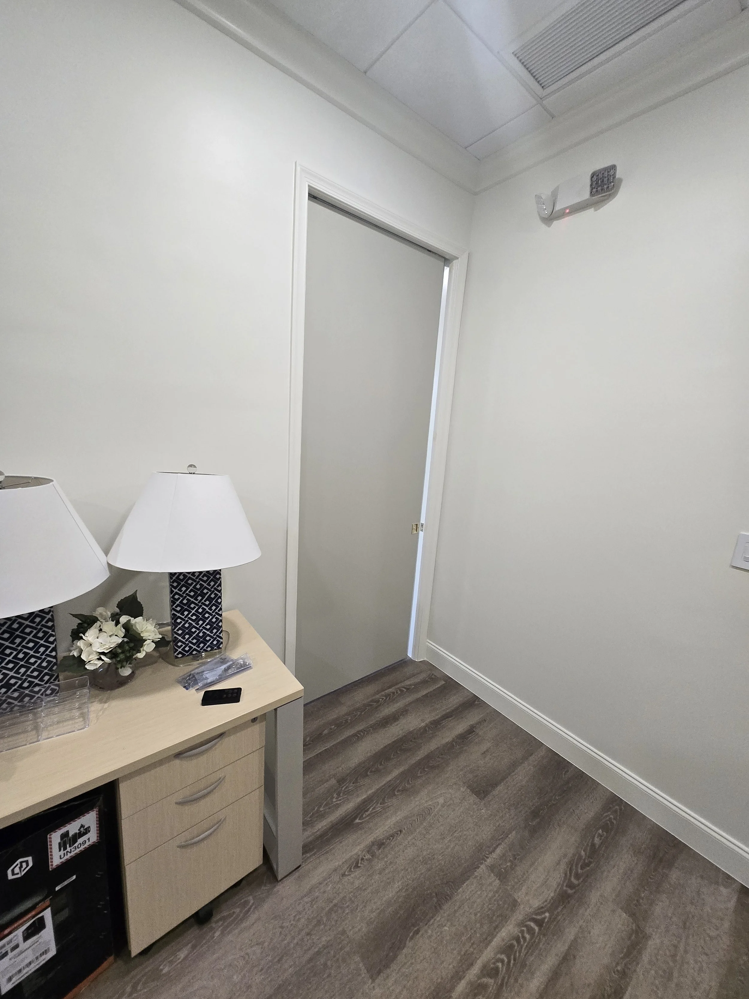 Empty room with white walls, wooden floor, a small table with lamps and decorative items, and a closed door.