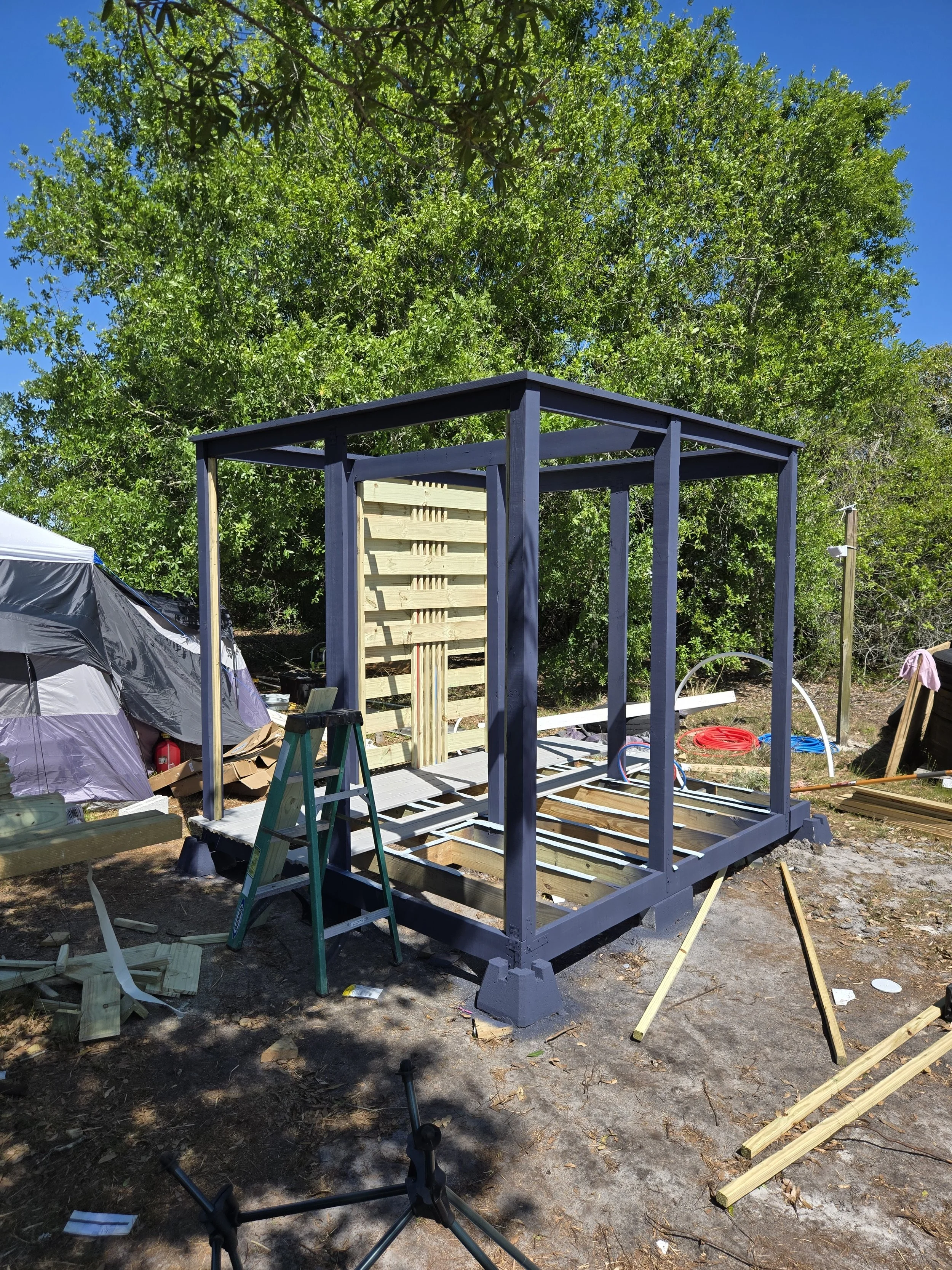 A small outdoor structure under construction with a wooden frame and partial wall, surrounded by tools, wood pieces, and construction materials, with trees and a blue sky in the background.