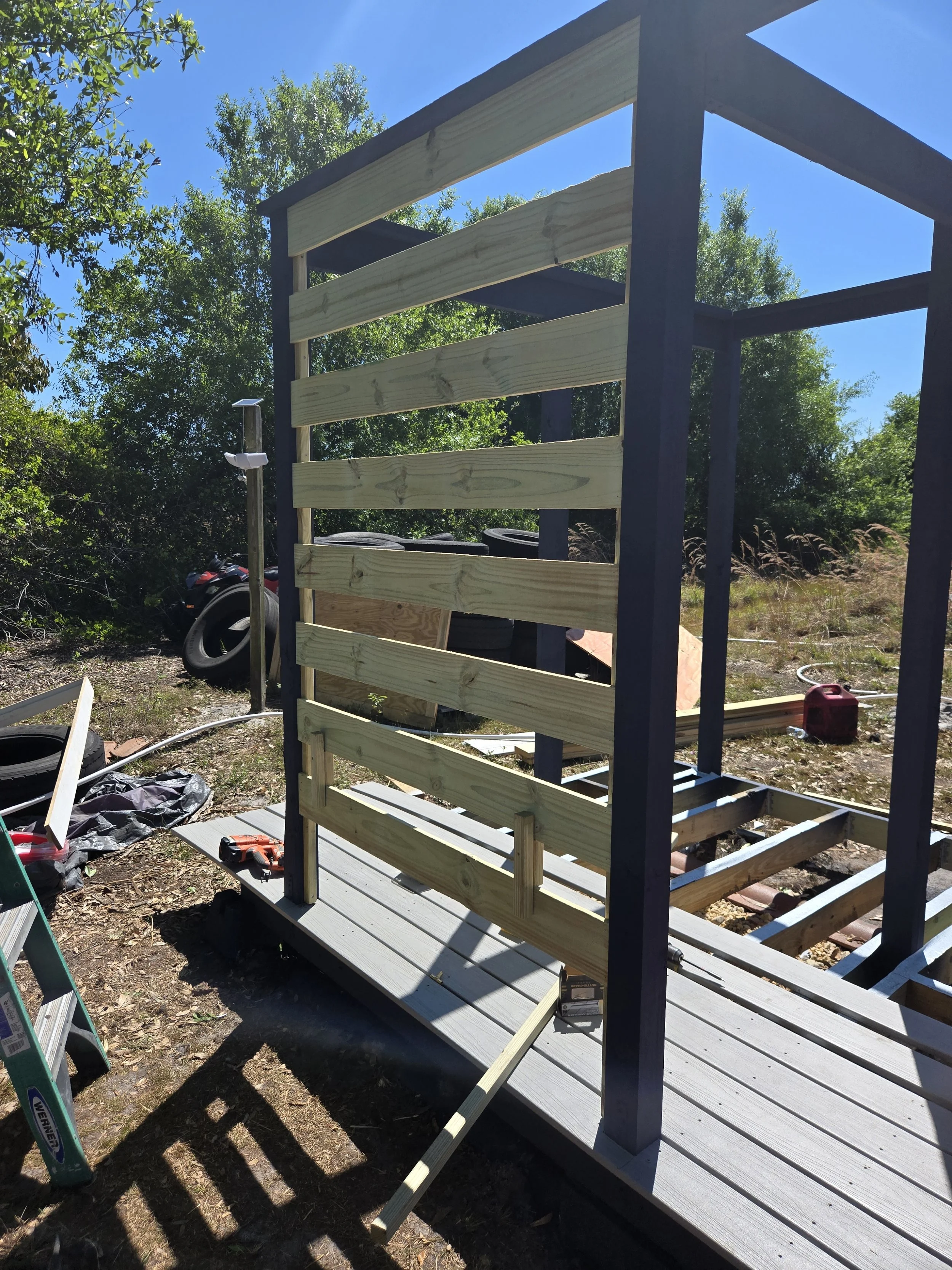 Construction of a wooden deck with vertical posts and horizontal slats on a sunny day, surrounded by trees and gardening tools.