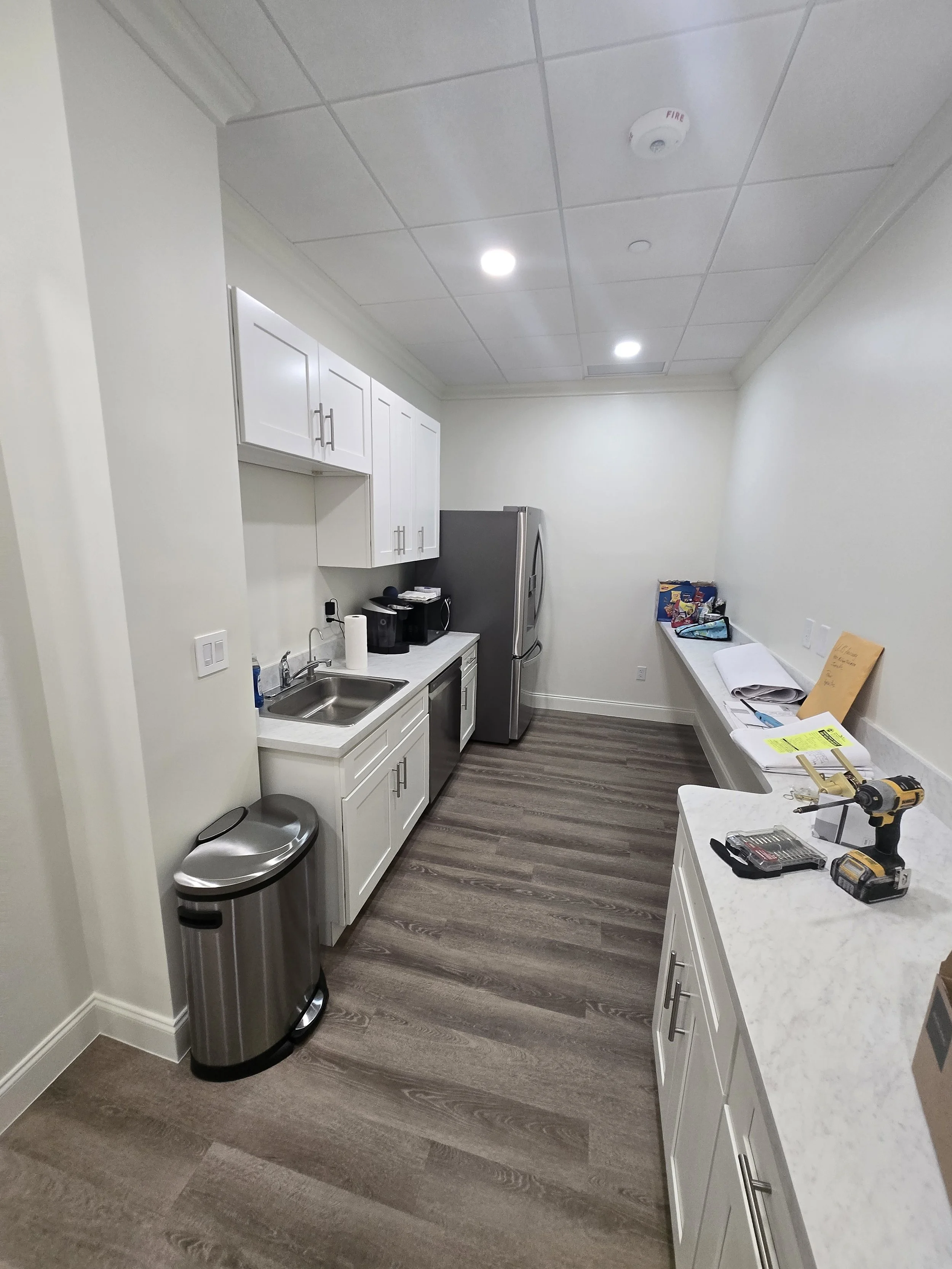 An empty kitchen or break room with white cabinets, a stainless steel refrigerator, a countertop with a coffee maker, sink, and various items, a trash can, and a long white counter with papers, a drill, and other tools.