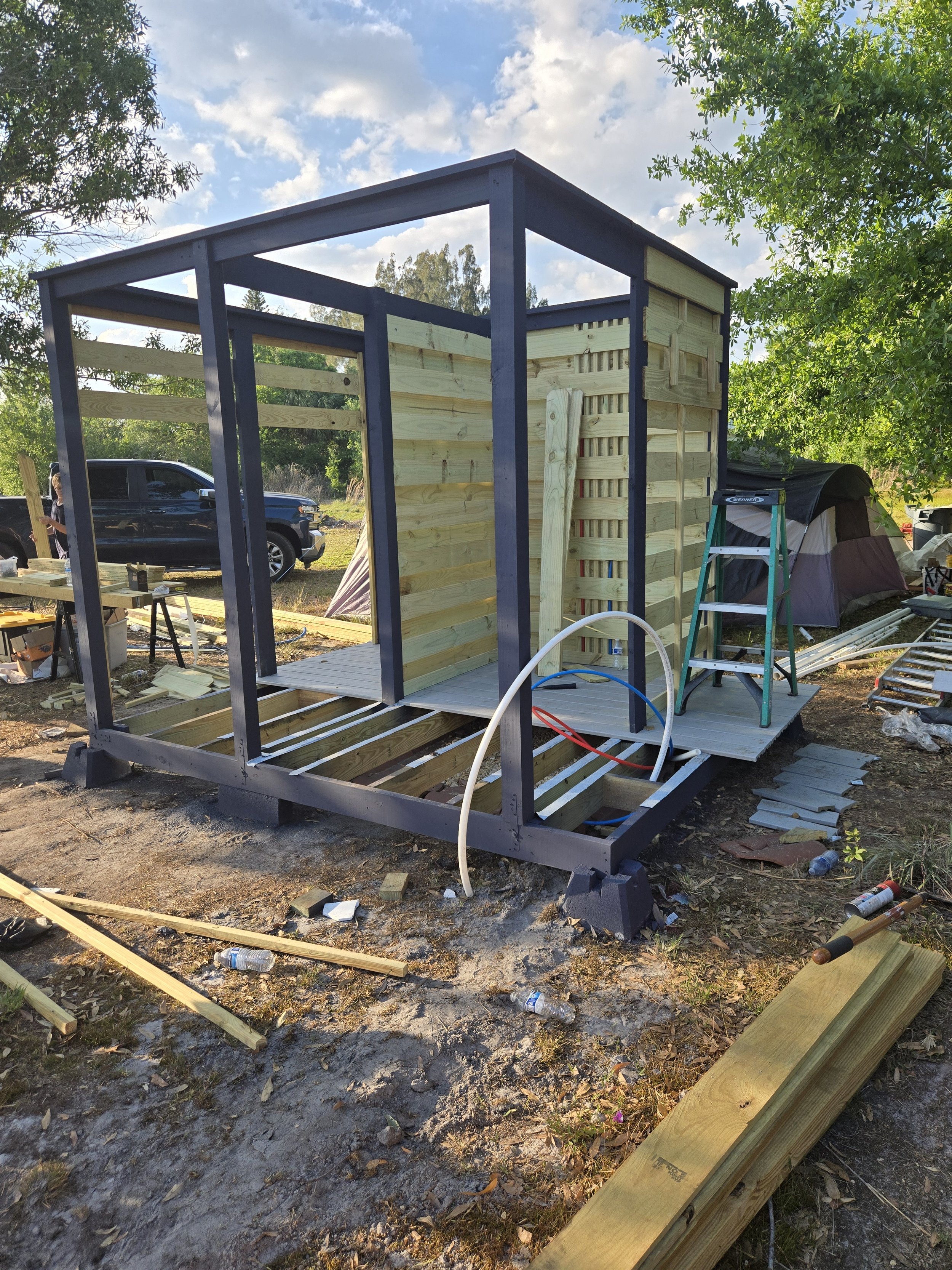 Construction of a small wooden shed outdoors with building materials, a ladder, and a black vehicle in the background.