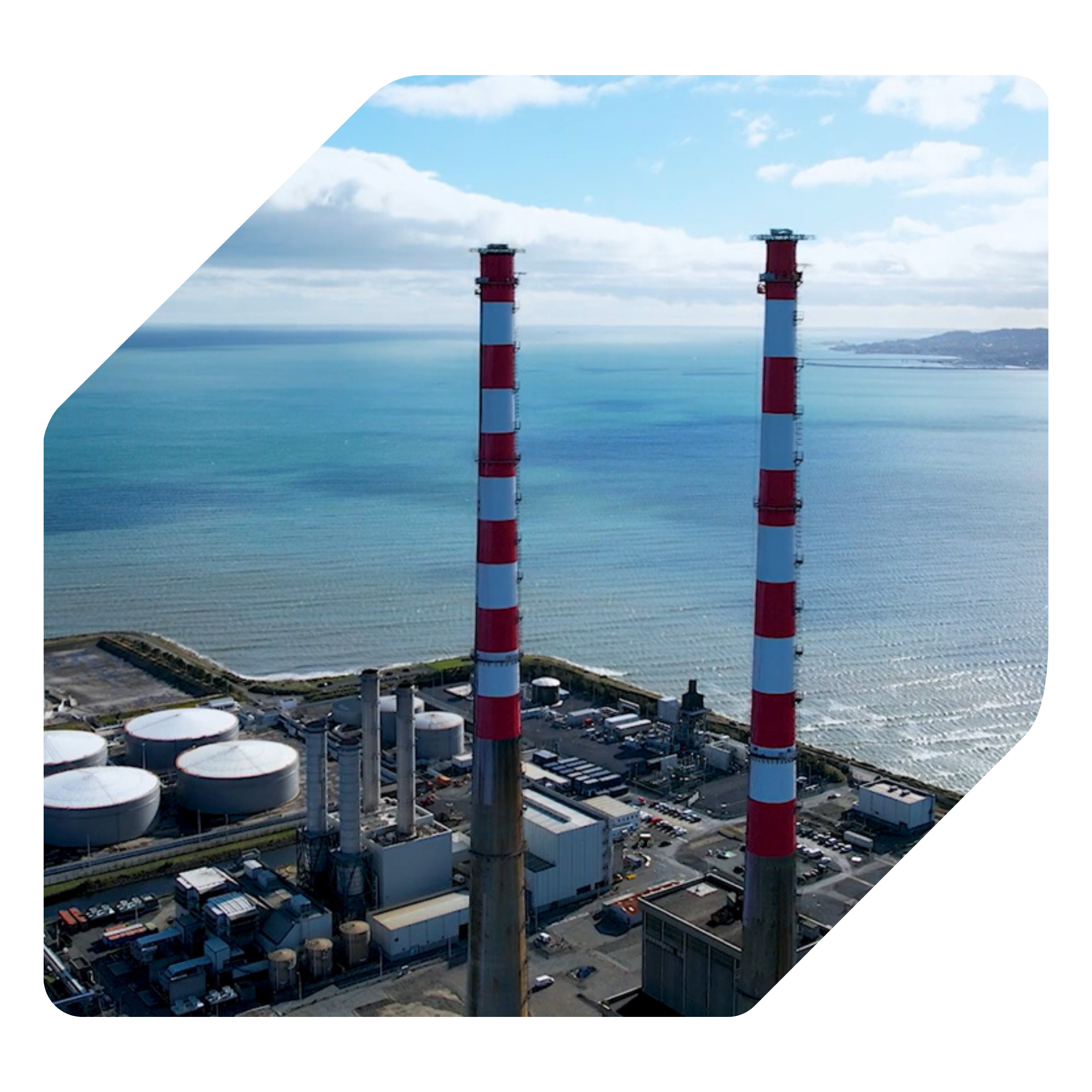 Industrial facility with two large red and white smokestacks near a body of water, surrounded by storage tanks and small buildings.