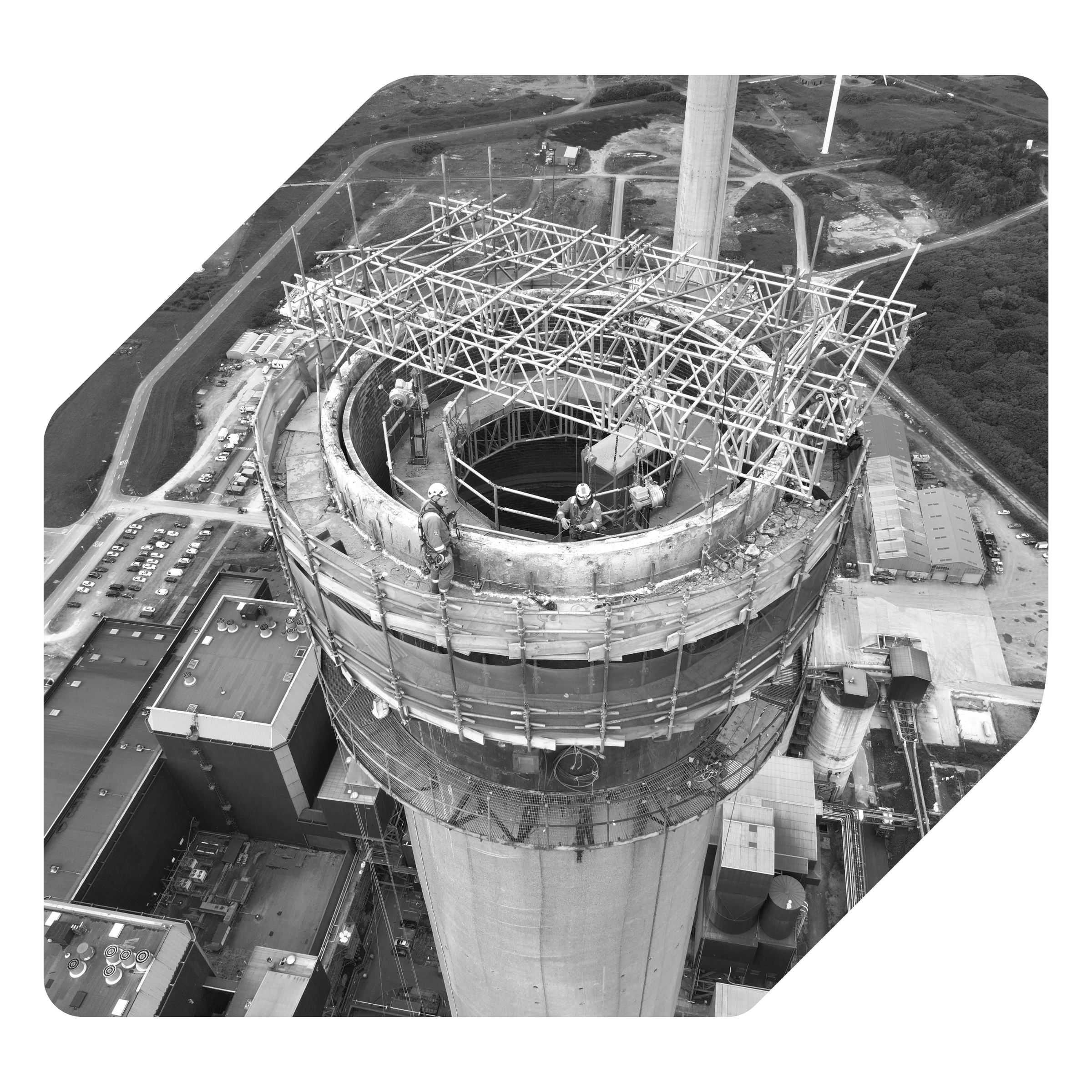 Aerial view of a large wind turbine construction site showing a tower and a complex steel structure at the top, with surrounding roads, buildings, and landscape in the background.