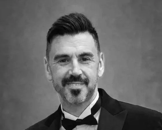 A black and white portrait of a man with short, dark hair styled to the side, a mustache and goatee, wearing a tuxedo with a bow tie, smiling at the camera.