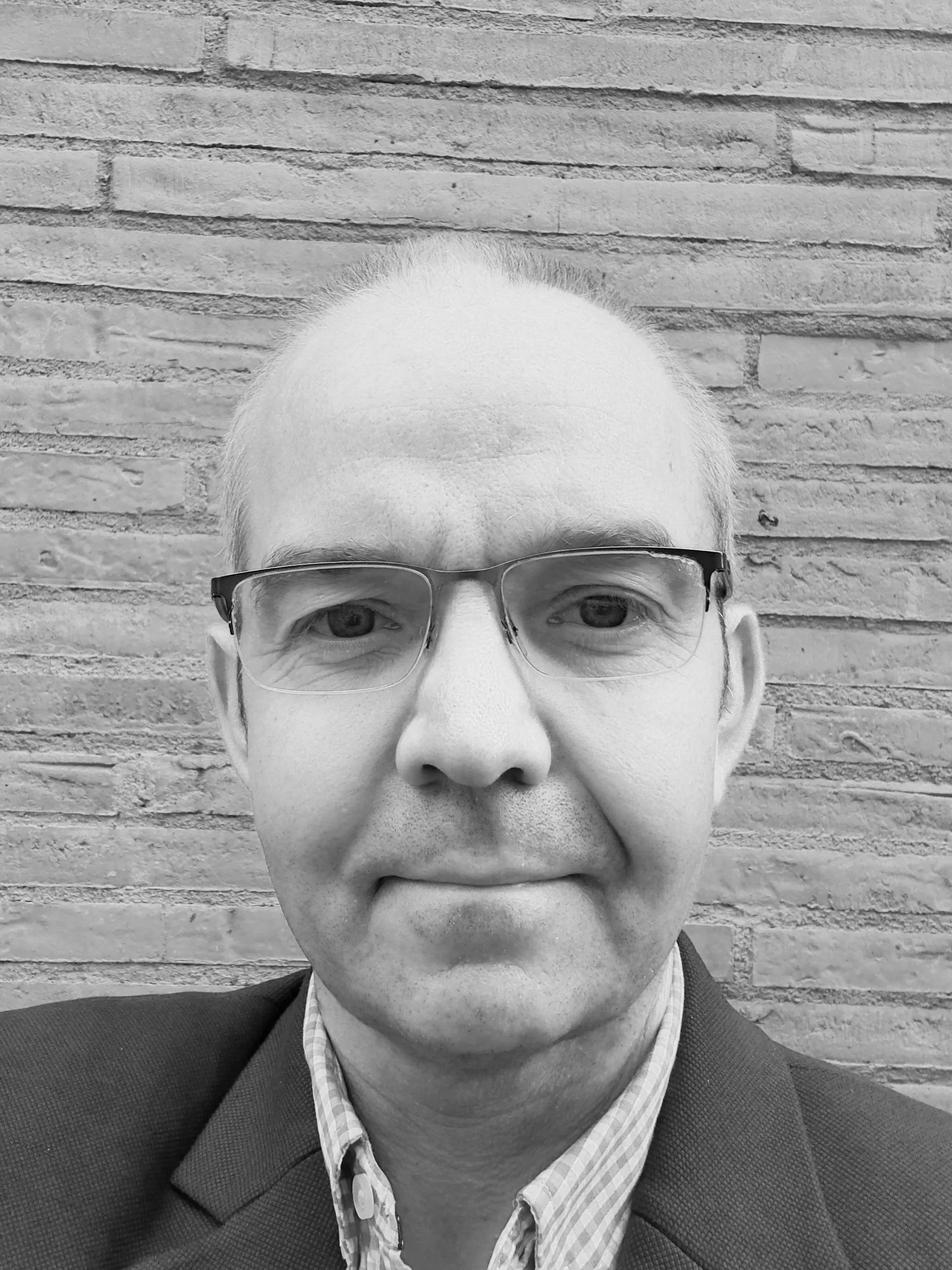 A black and white close-up of a man with short, light hair, wearing glasses, a shirt, and a blazer, standing against a brick wall background.
