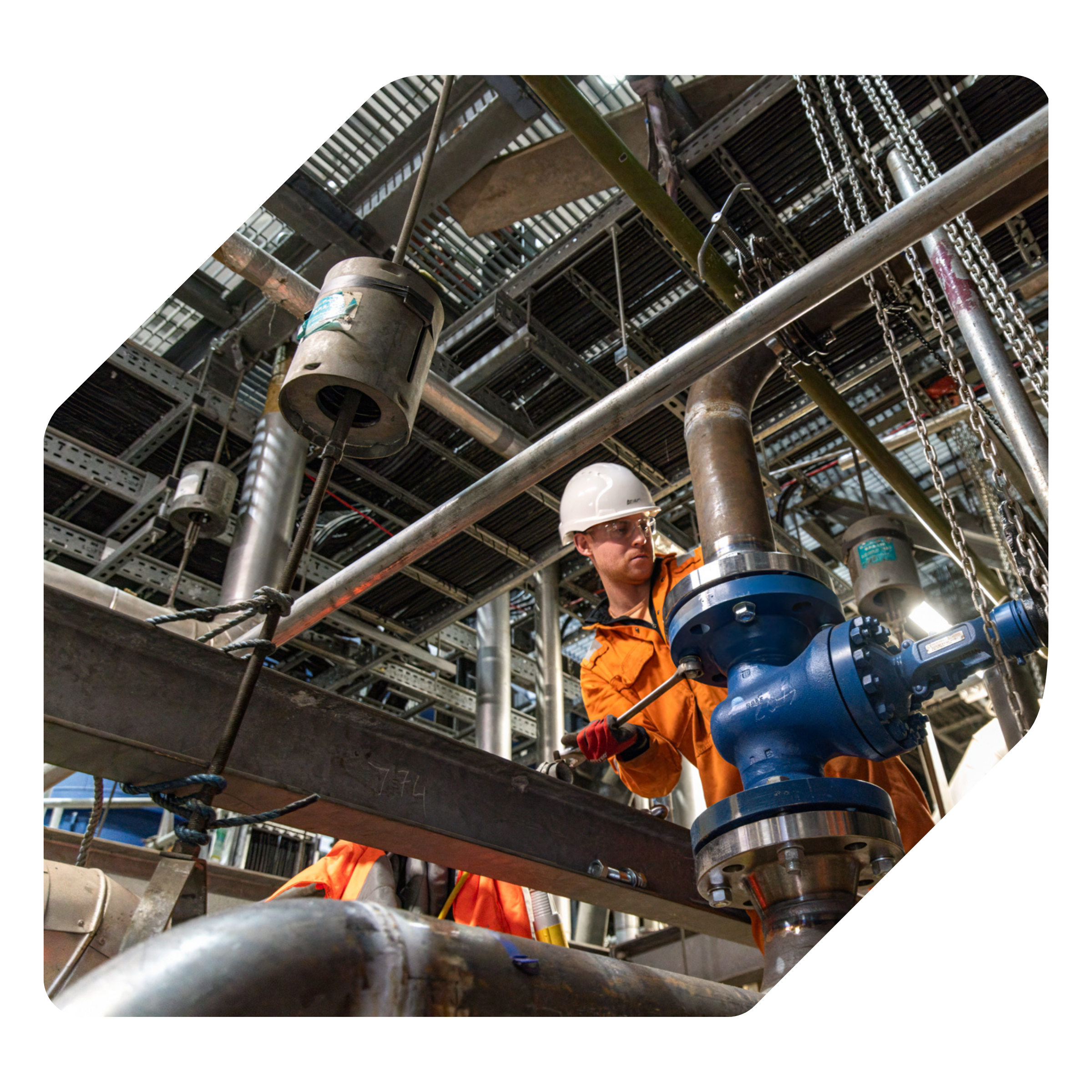 A construction worker wearing an orange jacket and a white helmet working with metal pipes and equipment on scaffolding at a construction site.