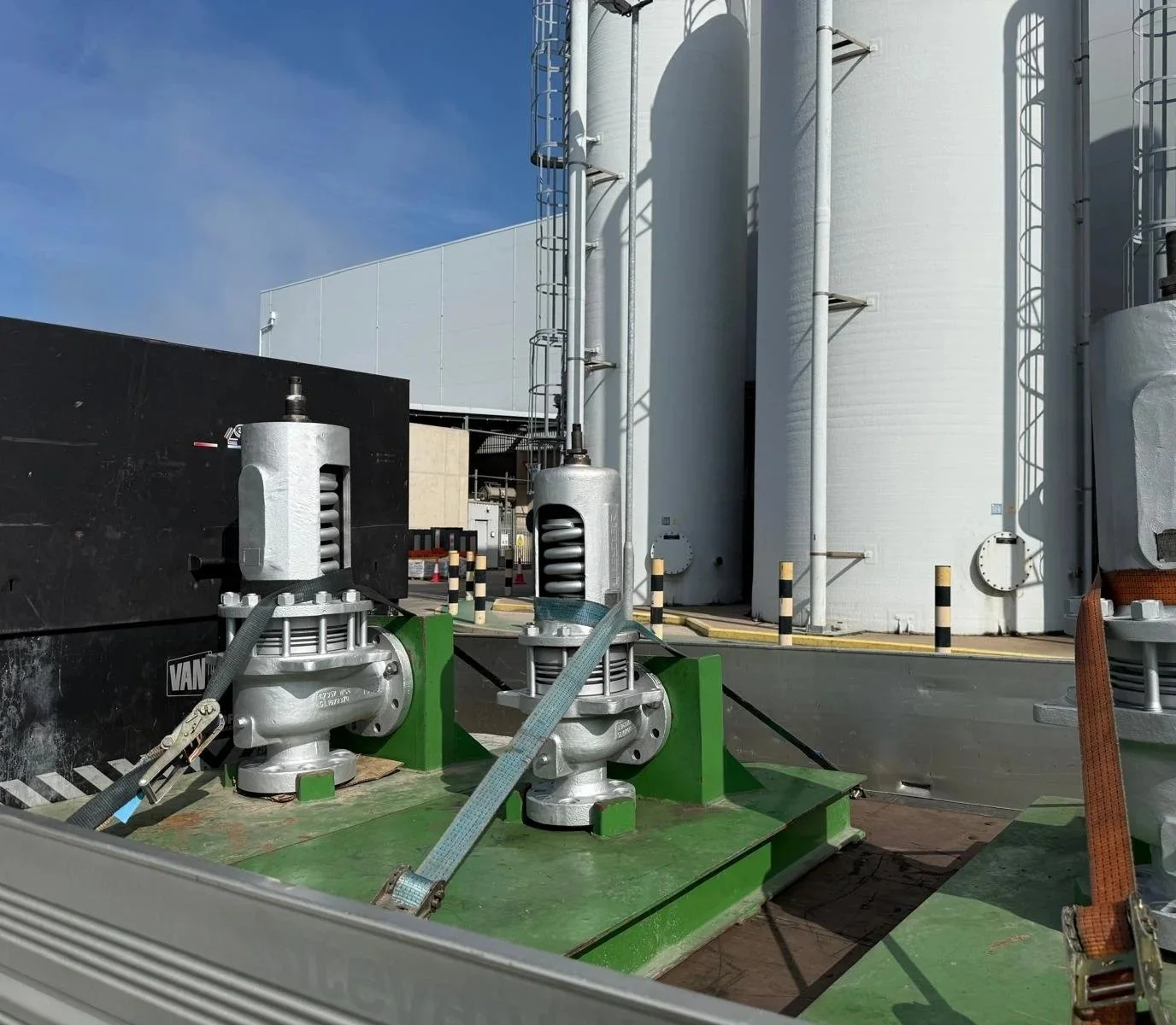 Close-up of industrial machinery on a rooftop, featuring large metal valves, pipes, and safety barriers near storage tanks under a clear blue sky.