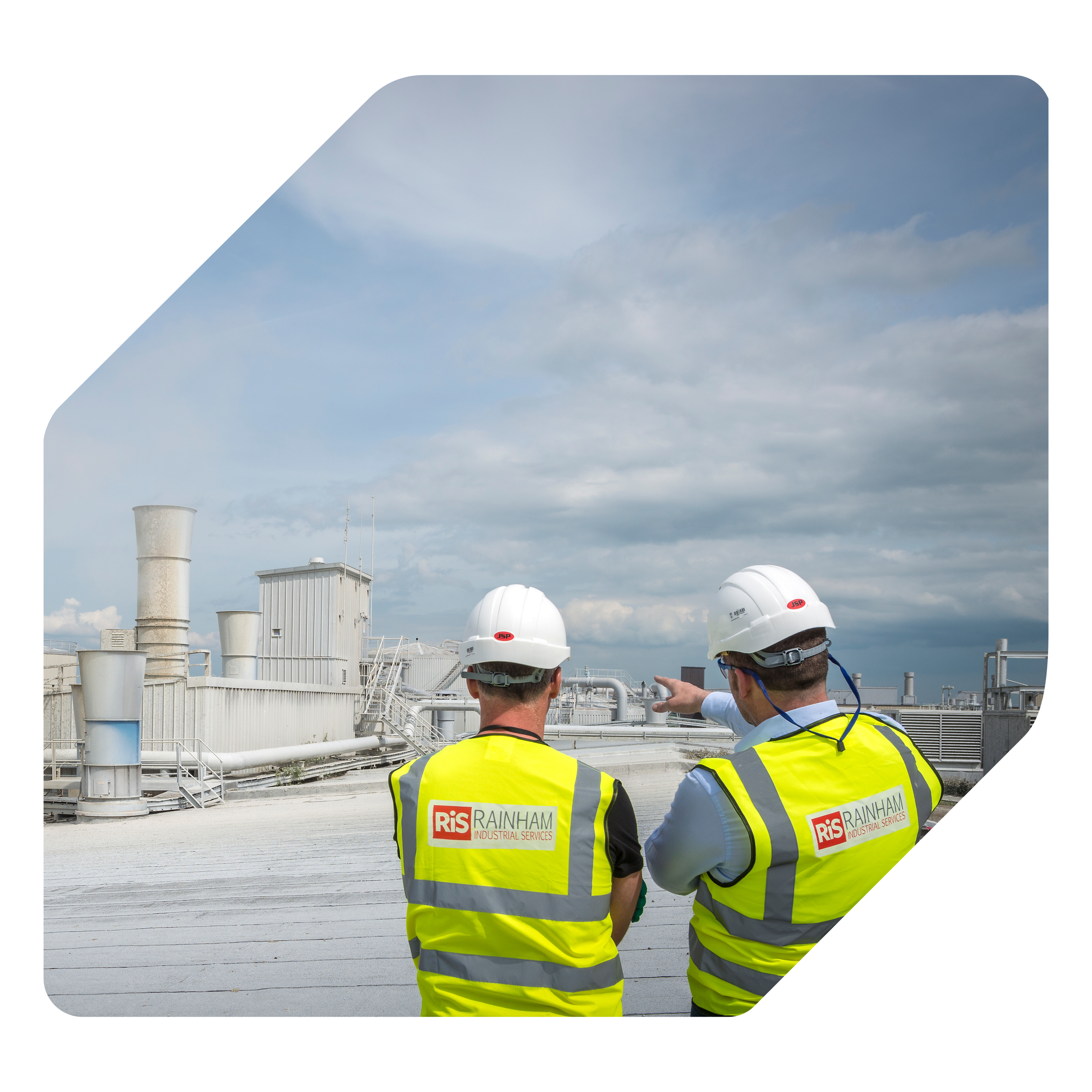 Two construction workers in yellow safety vests and white helmets are standing on a rooftop, overlooking industrial structures with pipes and ventilation systems under a partly cloudy sky.