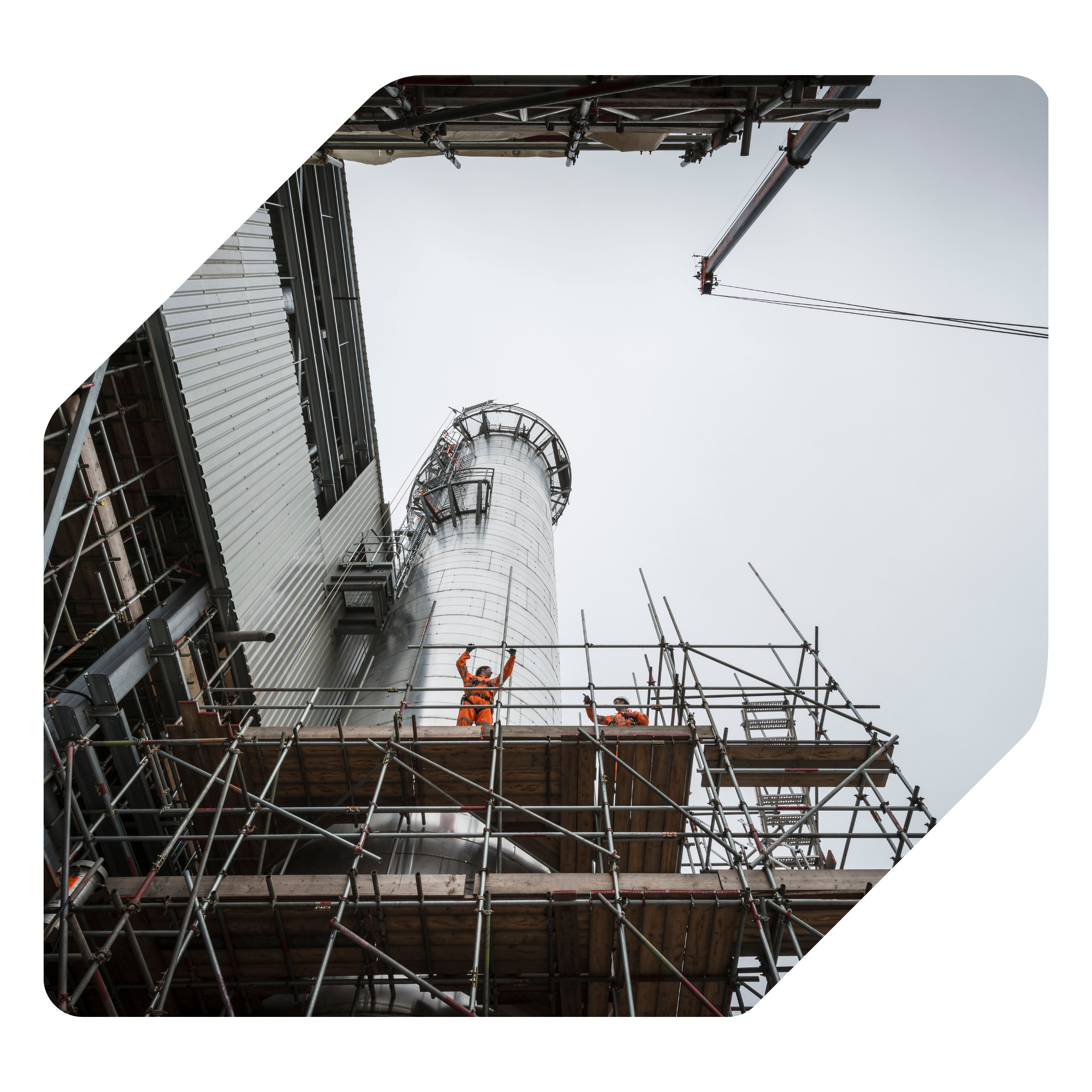 Workers in orange safety gear on scaffolding working on a large industrial chimney under a cloudy sky.
