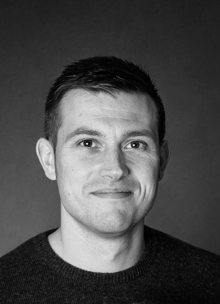 Black and white portrait of a man with short dark hair, wearing a dark sweater, smiling slightly, against a plain dark background.
