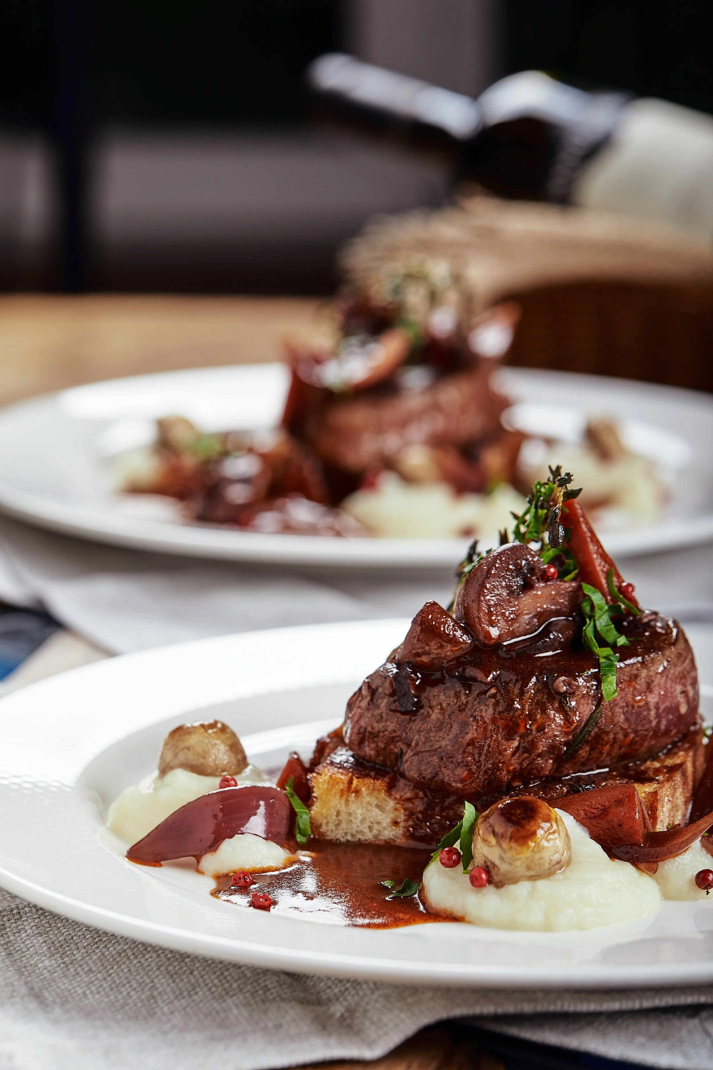 A plated serving of gourmet beef steak with mashed potatoes, garnished with herbs, mushrooms, and a rich sauce, set on a white plate on a table.