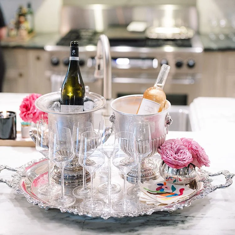 Champagne bottles chilling in ice buckets on a silver tray with empty champagne flutes, pink flowers, and decorative napkins on a white kitchen counter.