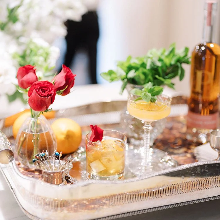 A silver tray with a small vase of red roses, two yellow peaches, two cocktails with mint and flower garnishes, and decorative jewelry, all displayed on a table with blurred background.