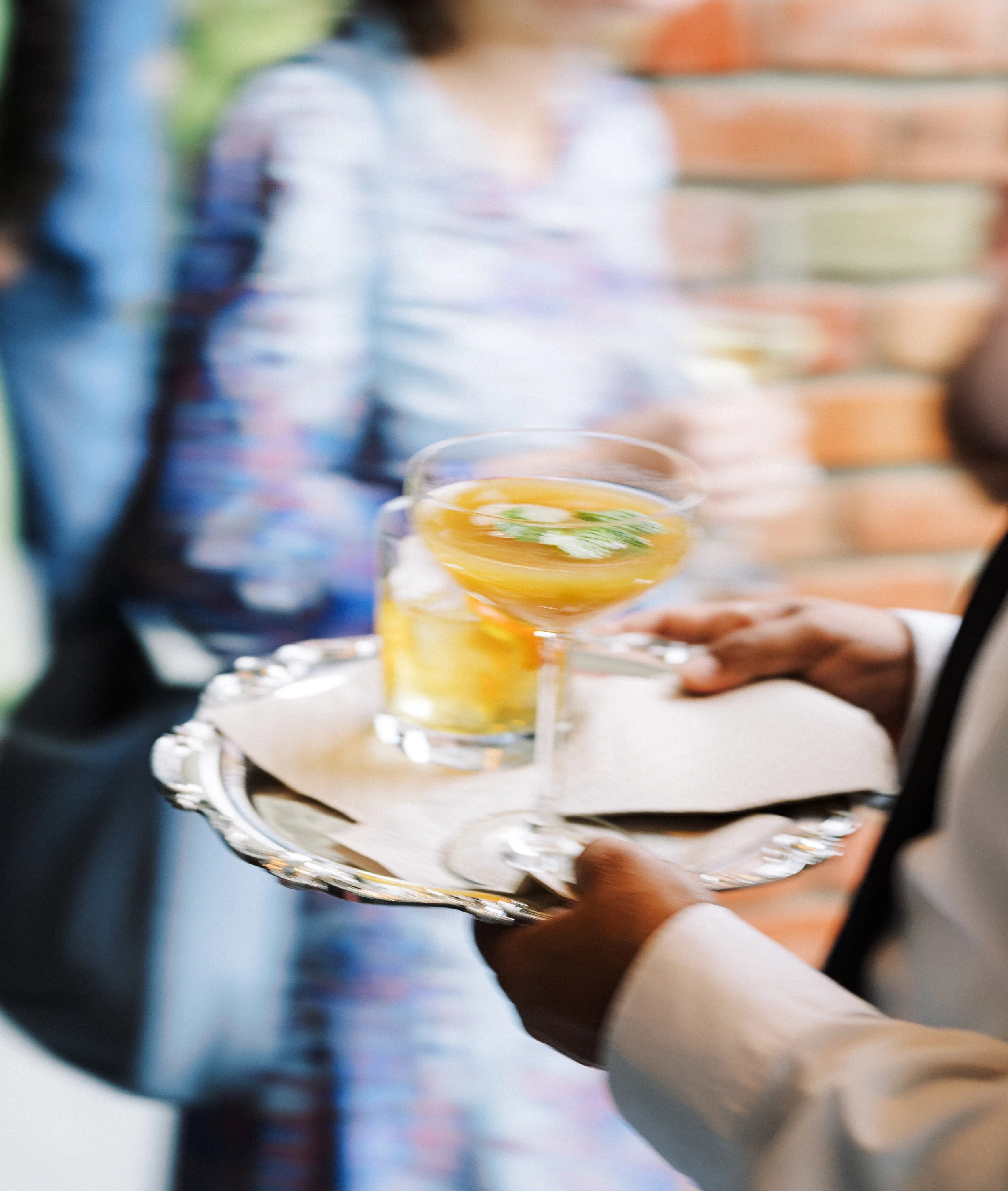 A person in a suit holding a silver tray with a glass of yellow cocktail garnished with mint leaves and slices of citrus, during an event or gathering.