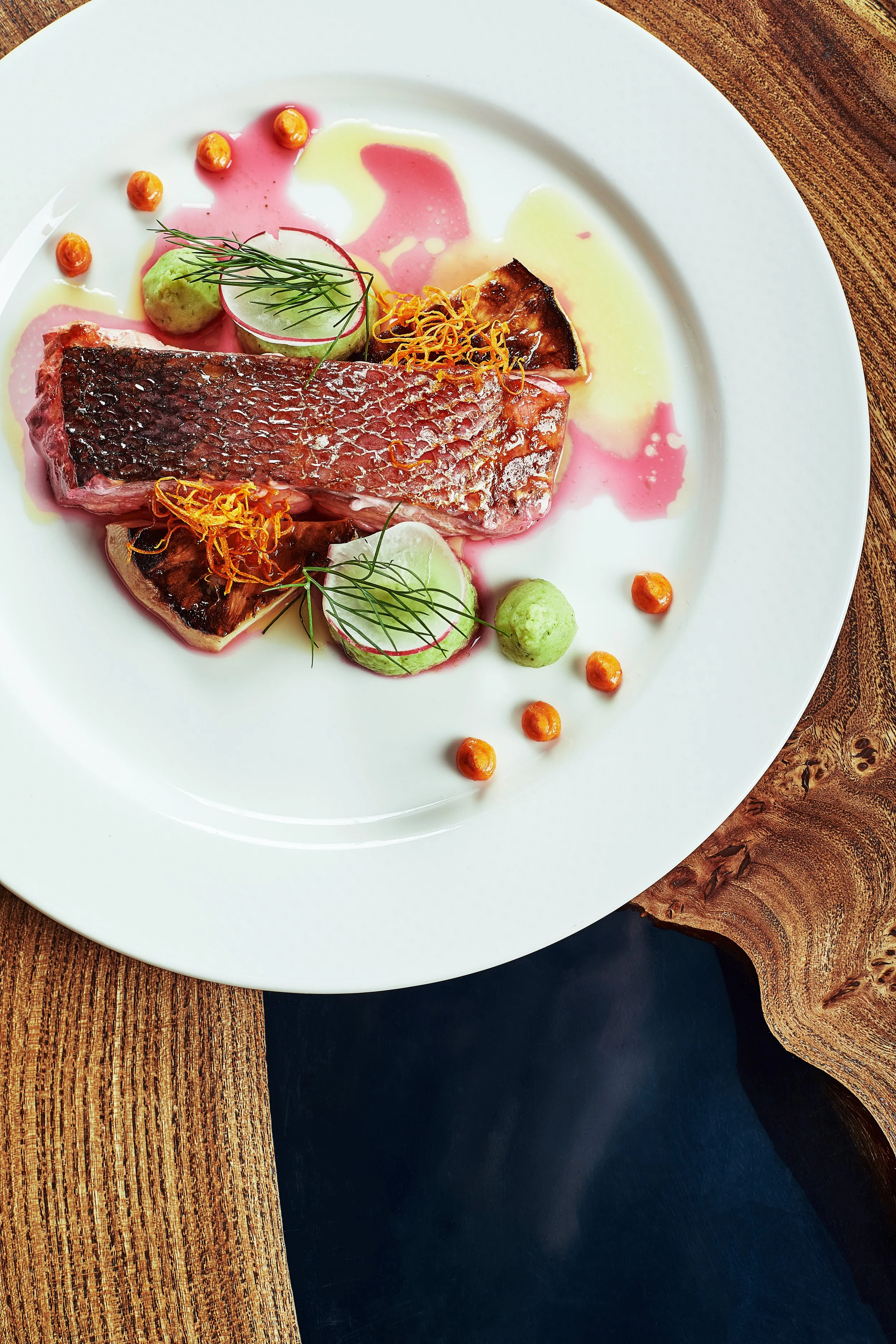 Gourmet plated dish with a portion of seared fish, garnished with microgreens, roasted vegetables, dollops of green and orange sauces, and colorful edible sprigs, on a white plate on a wooden table.