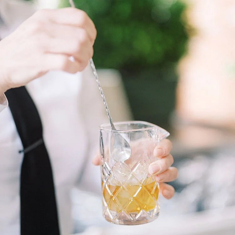 A person stirring a cocktail in a glass with ice, garnished with lemongrass or a similar herb