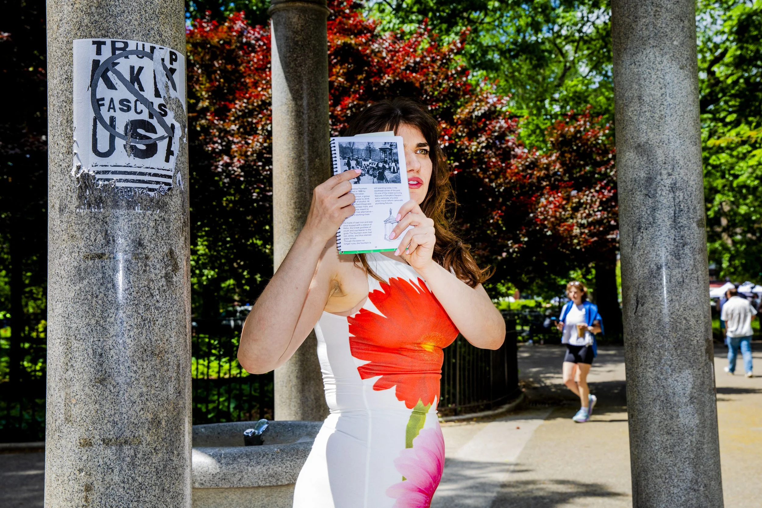 Woman in a white dress with floral patterns reading a booklet in a park with trees and people walking in the background.