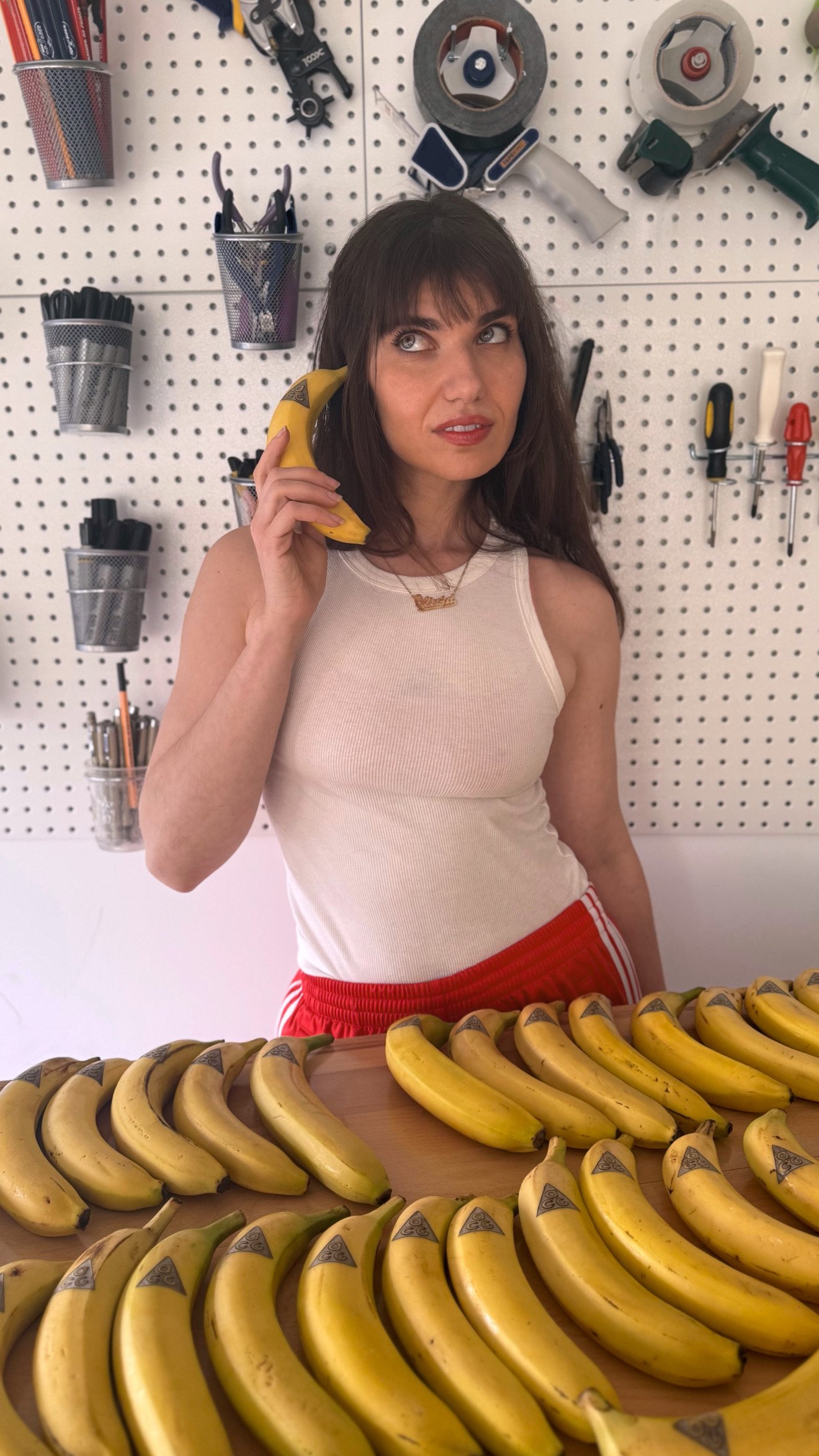Woman with brown hair and bangs talking on a banana-shaped phone, standing behind a table full of bananas, with a wall of tools and supplies in the background.